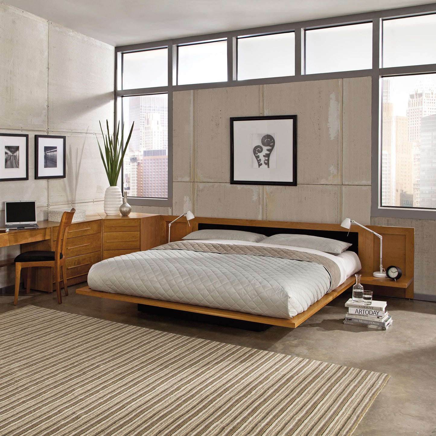 Modern bedroom featuring the Copeland Furniture Moduluxe Platform Bed with Upholstered Headboard - 29" Series, adorned with white bedding. A painting and two small white lamps are mounted above its wooden headboard. To the left, a wooden desk accompanied by a chair and potted plant adds to the room's charm. Natural light streams through large windows, illuminating a striped rug on the floor.