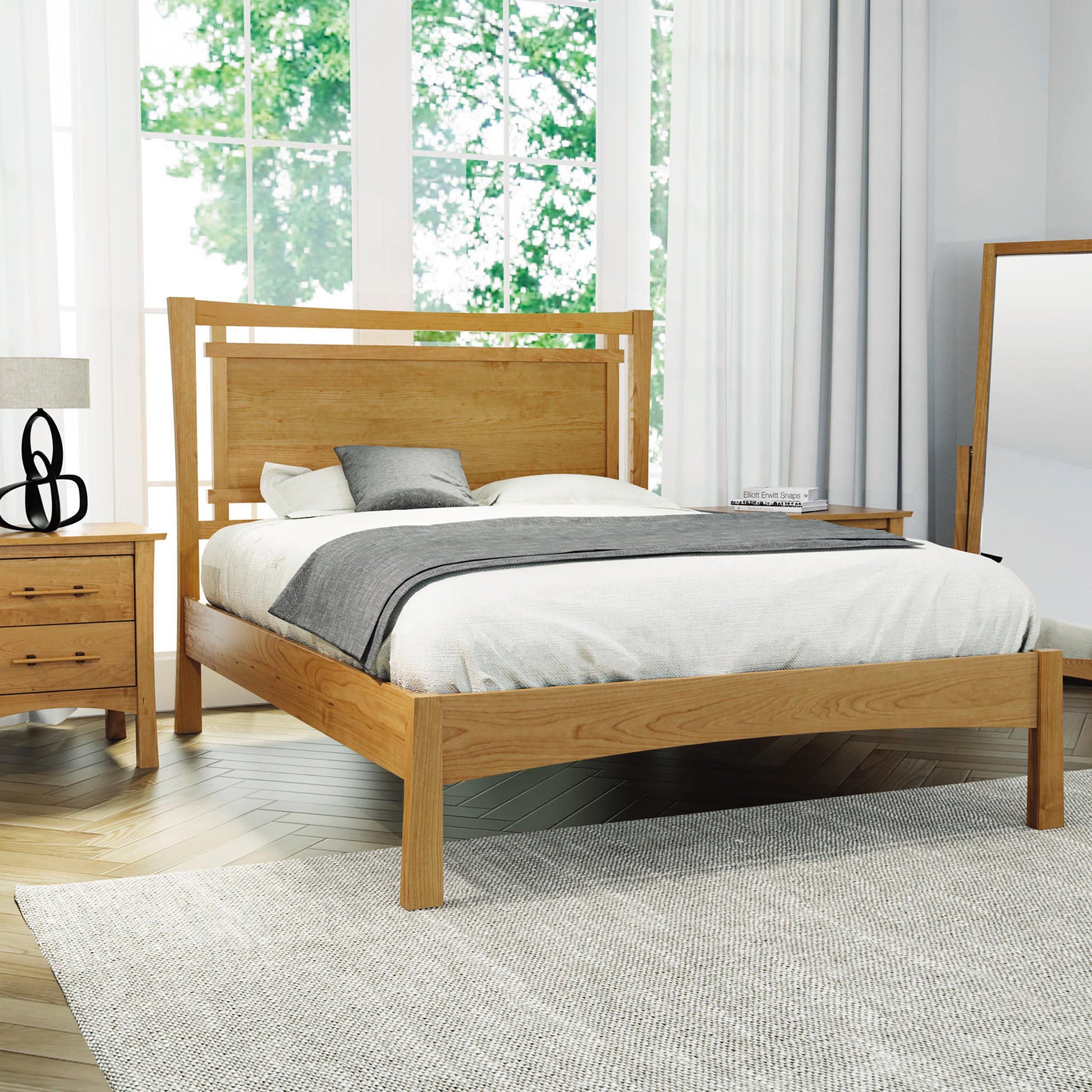 A bedroom features the eco-friendly Monterey Platform Bed by Copeland Furniture, crafted from solid cherry wood, adorned with a white and gray bedspread. To the left stands a matching wood nightstand with drawers, topped by a lamp and a decorative item. A large window dressed in white curtains lets in natural light, while to the right there's a full-length mirror. The floor is covered by a light-colored rug.