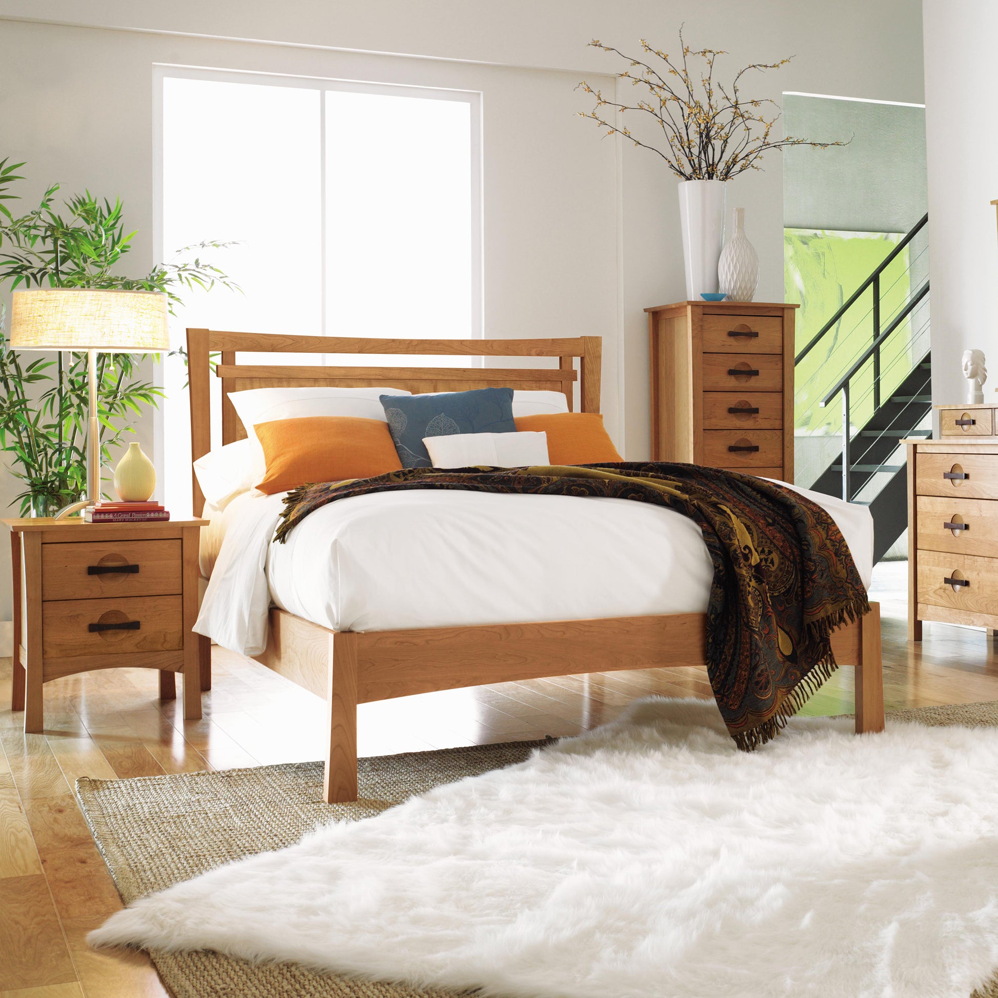 A bedroom featuring a solid cherry wood Monterey Platform Bed from Copeland Furniture, complemented by matching nightstands and a dresser. This eco-friendly bed is adorned with white and orange bedding, topped with a patterned blanket. Additionally, the nightstands hold a lamp and plant each, while the dresser displays a vase with branches. A large white rug covers the floor.
