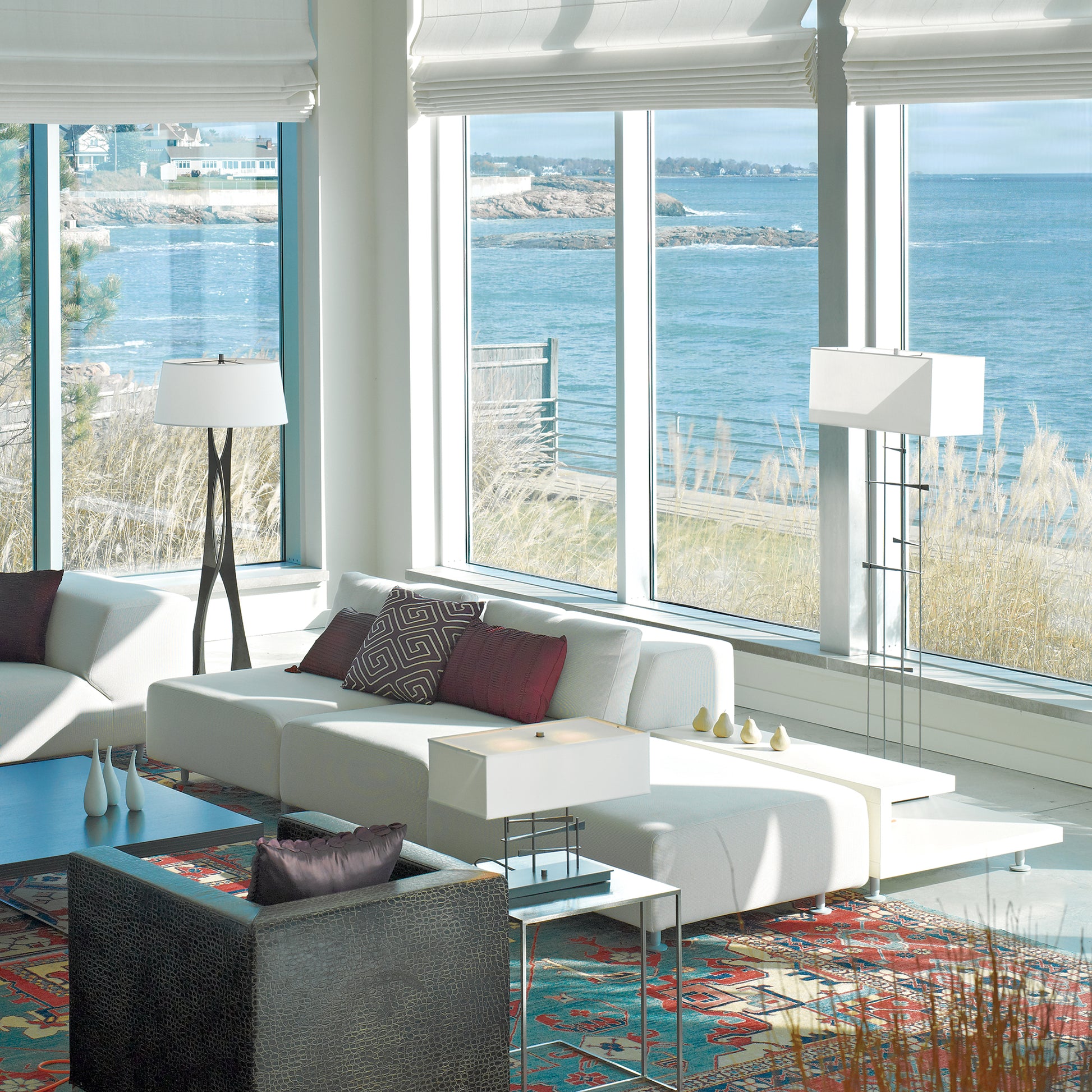 Modern, bright living room with large windows offering a view of the ocean. The room features a white sofa, stylish Hubbardton Forge Moreau Floor Lamp, and a colorful rug on a white floor.