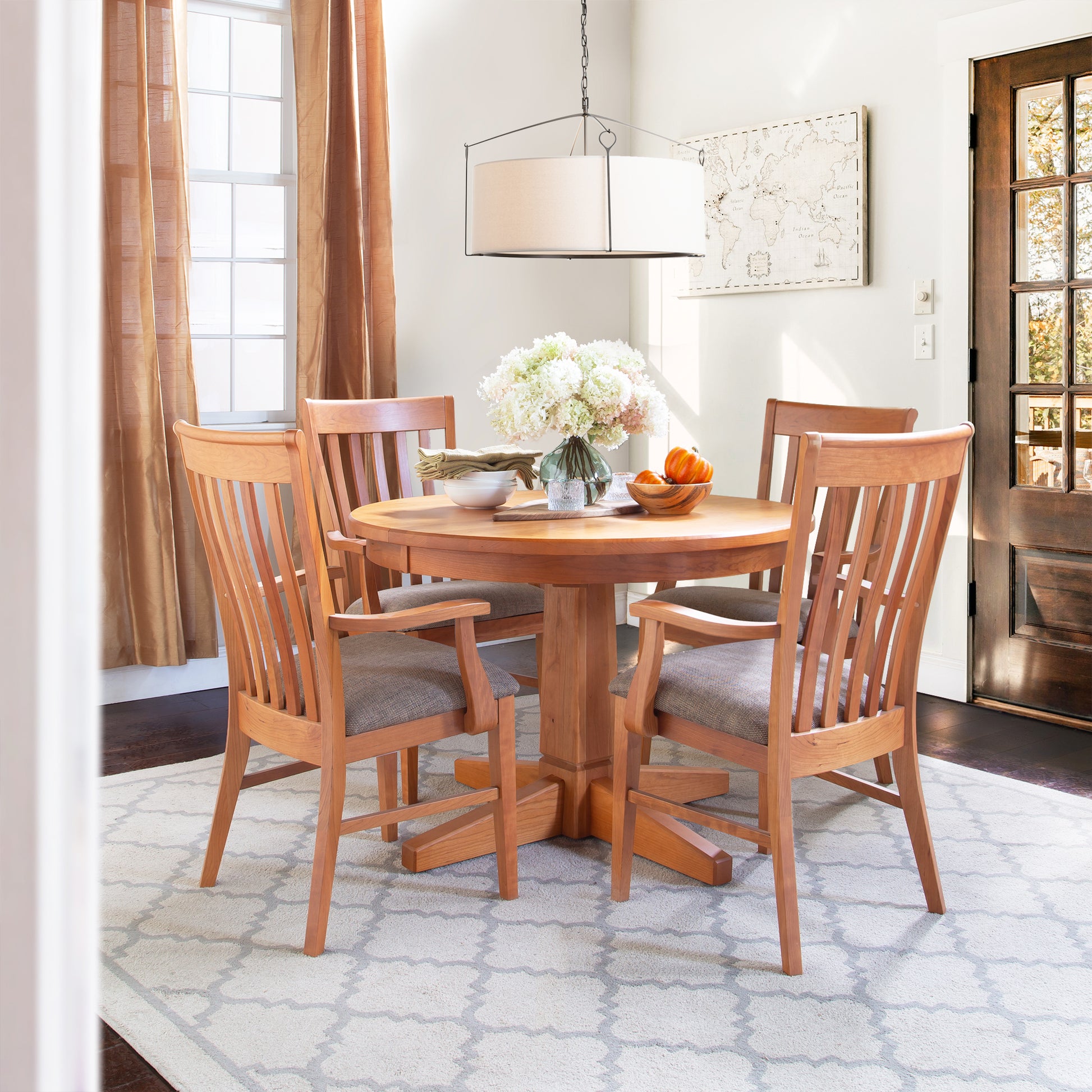 A round handcrafted Natural Vermont Single Pedestal Round Solid Top Table from Lyndon Furniture, accompanied by four matching chairs, is set in a bright room with a large window. The table features a vase of white flowers and a bowl of fruit. A pendant light hangs above, and framed artwork adorns the wall. This eco-dining room furniture piece is elegantly placed on a geometric rug.