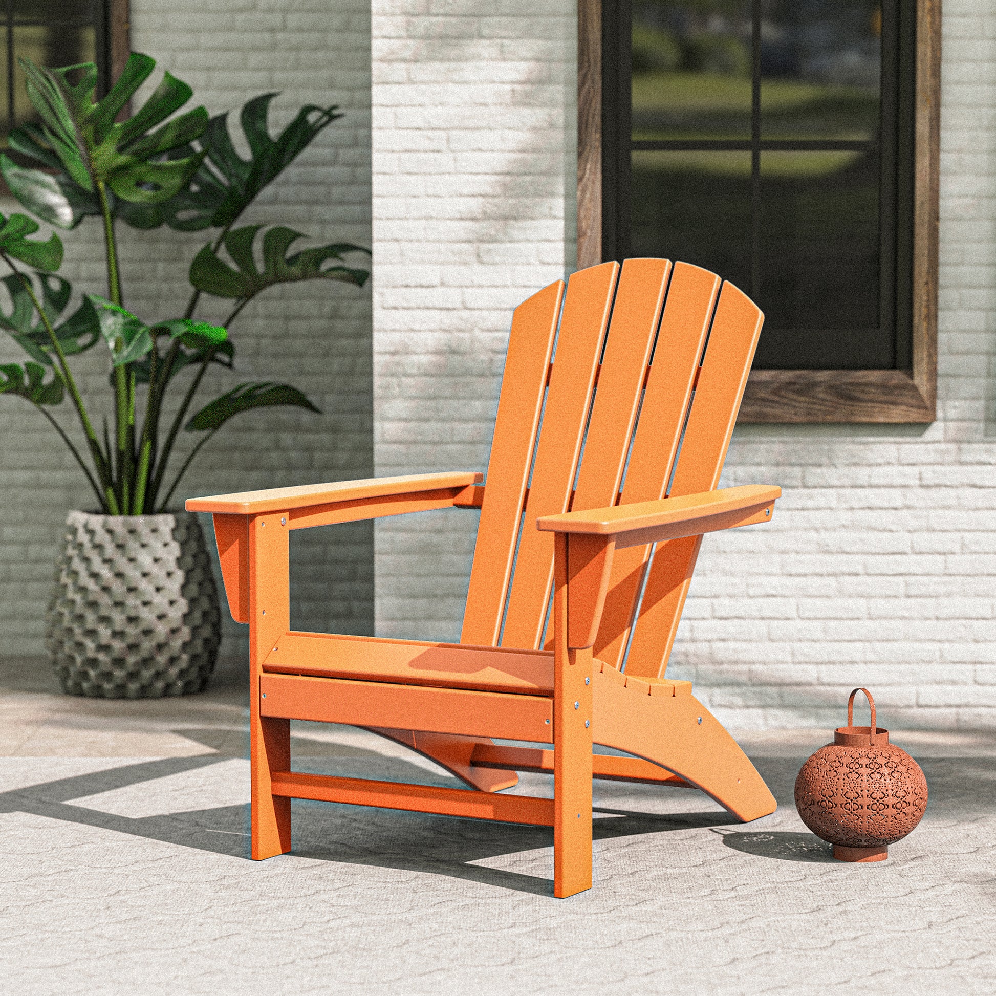 An orange POLYWOOD Nautical Adirondack Chair is placed on a patio with a light gray surface, set against a white brick wall. To the left, there's a potted plant, and in the background, you can see a window with dark trim. Nearby, a small round lantern adds to this perfect outdoor relaxation spot.