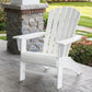 A POLYWOOD Nautical Curveback Adirondack Chair in a sleek white finish, made from robust POLYWOOD lumber, is positioned on a stone patio near a brick column, offering an ideal spot for outdoor relaxation. The scene is complemented by a vibrant green lawn and flourishing shrubs in the background.