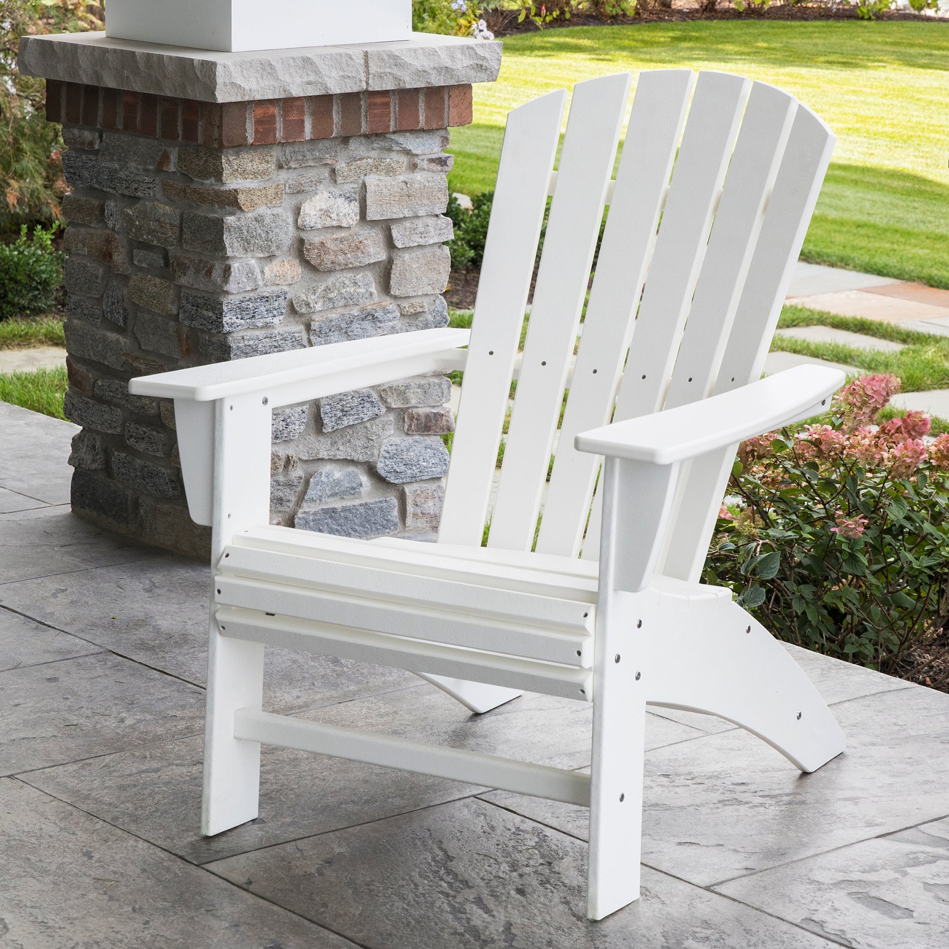 A POLYWOOD Nautical Curveback Adirondack Chair in a sleek white finish, made from robust POLYWOOD lumber, is positioned on a stone patio near a brick column, offering an ideal spot for outdoor relaxation. The scene is complemented by a vibrant green lawn and flourishing shrubs in the background.