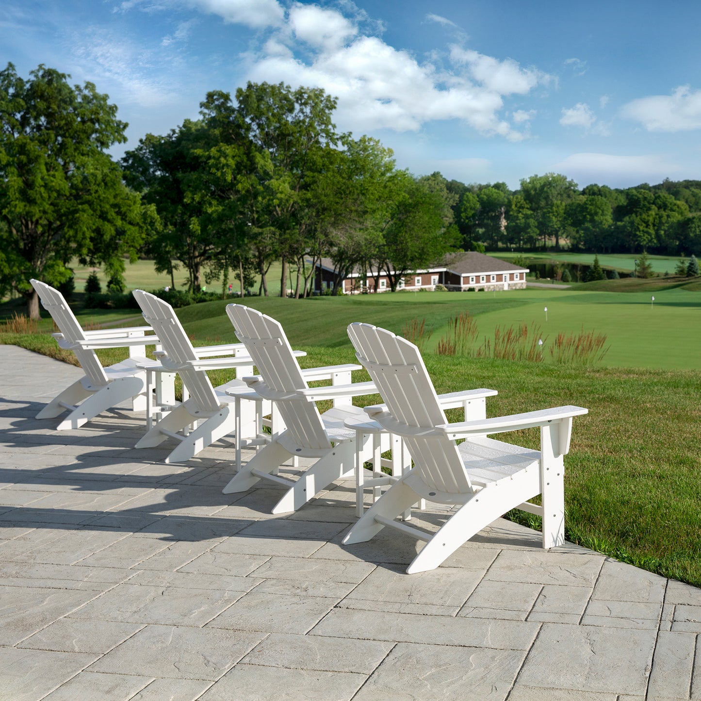 Four POLYWOOD Nautical Curveback Adirondack Chairs, made from sturdy POLYWOOD lumber and finished in white, are set on a stone patio that overlooks a beautifully maintained golf course. The outdoor relaxation area is surrounded by trees under a partly cloudy sky, with the clubhouse visible in the background.