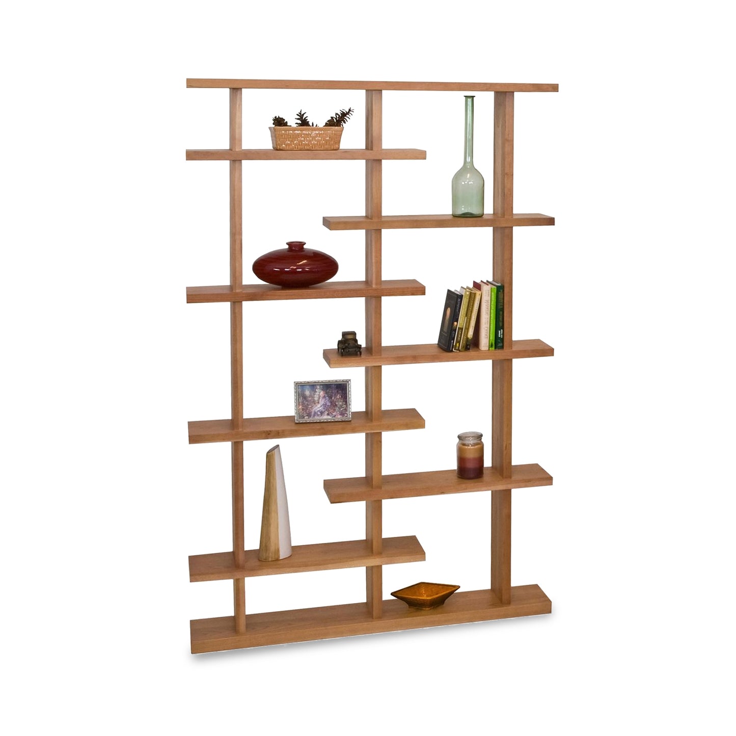 The New York Contemporary Bookcase by Lyndon Furniture showcases American-made, solid wood craftsmanship with its staggered shelving design. This modern bookcase features cherry wood and walnut wood shelves displaying a variety of decor: a potted plant, vase, photo frame, books, bottle, red ceramic piece, candle, and wooden tray. Perfect for those seeking high-quality Vermont made furniture for their home office or living room.