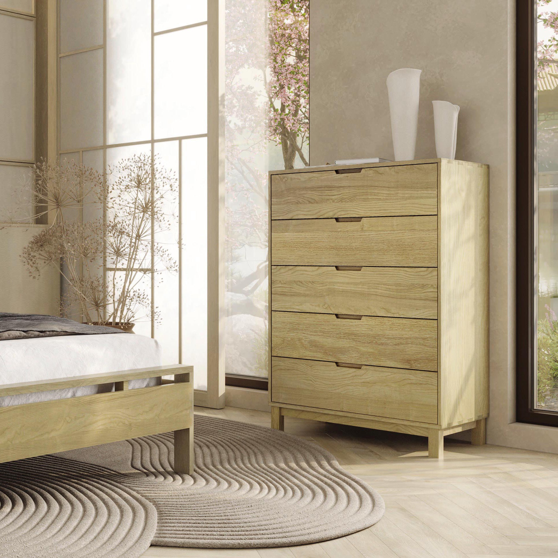 A modern bedroom with Solid Oak Hardwood Copeland Furniture Oslo Bedroom Furniture Collection, featuring a bed and an Oslo 5-Drawer Wide Chest, accentuated by a gray circular rug and tall white vases; natural light
