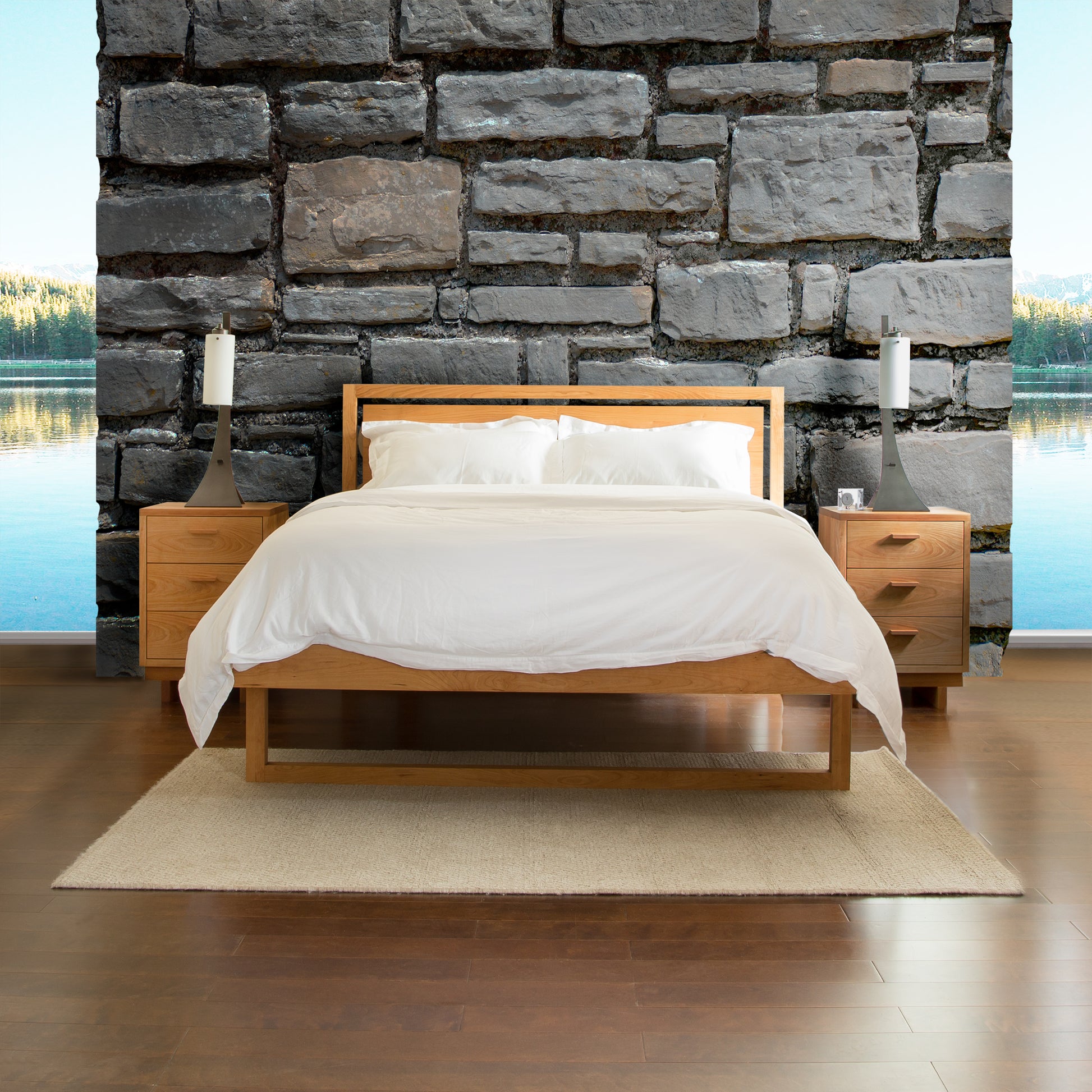 A modern Vermont Furniture Designs Pendant Bed made from sustainably harvested cherry wood with white bedding in front of a stone wall, overlooking a serene lake through a large window.