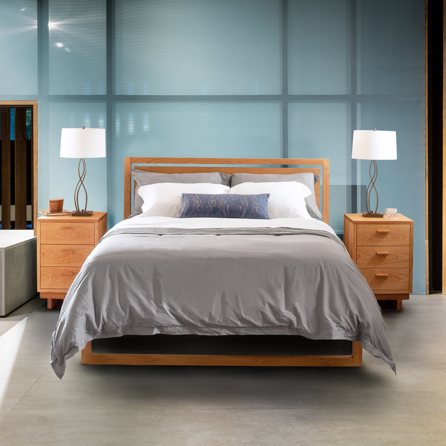 A neatly made Vermont Furniture Designs Pendant Bed with white and gray bedding, flanked by two wooden nightstands with matching lamps, set against a blue glass background.