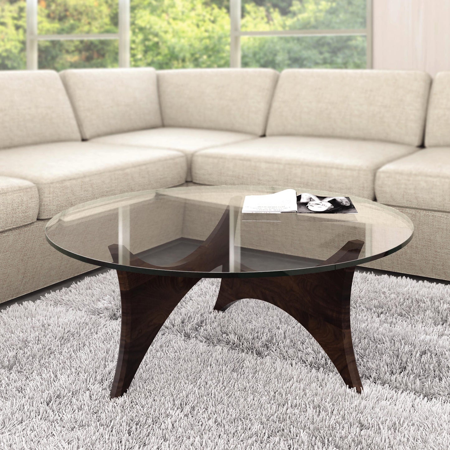 In a modern living room, the centerpiece is a Pivot Round Coffee Table by Copeland Furniture, featuring a tempered glass top and walnut base, gracefully set on a light gray shaggy rug. Complementing this chic setup is an L-shaped arrangement of a light beige sectional sofa around the table. On the coffee table rest a magazine and a remote. Large windows in the background flood the room with natural light and offer views of lush greenery.