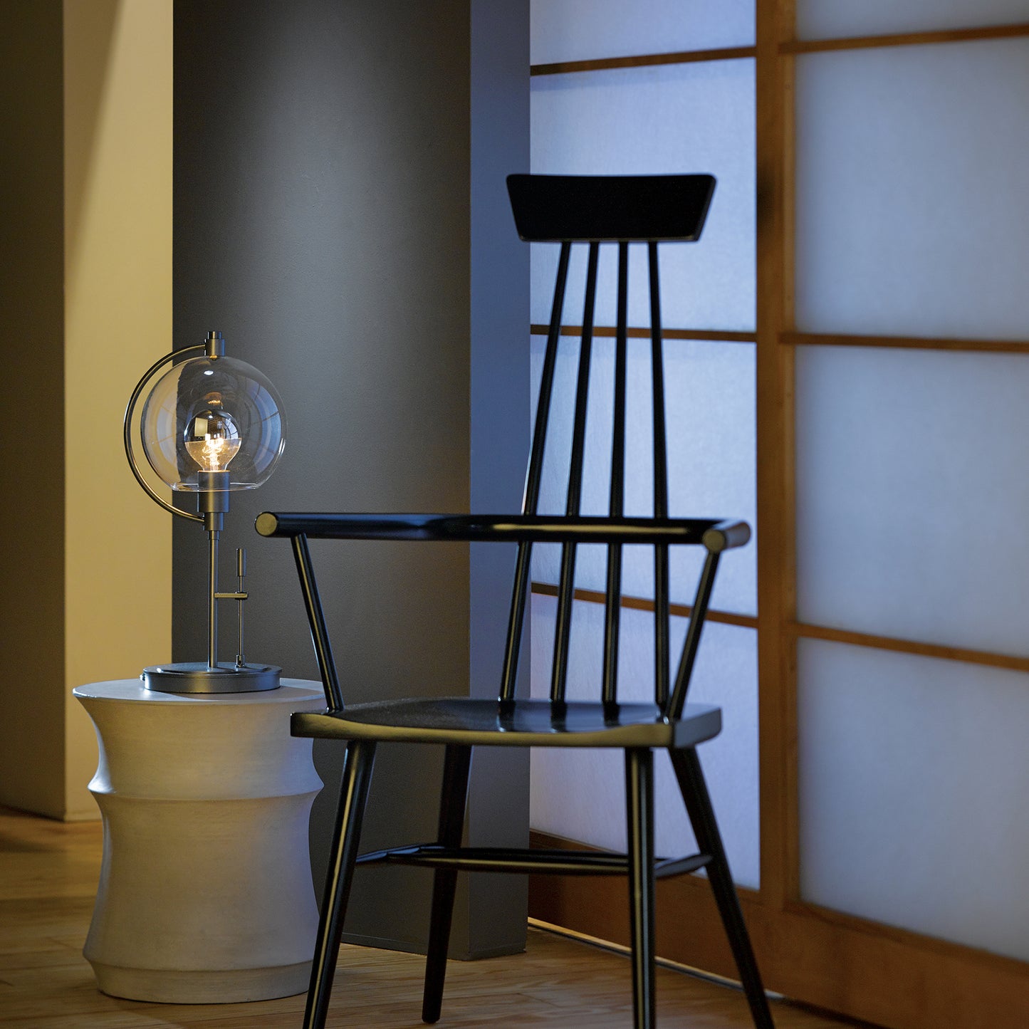 A black chair next to a Pluto Table Lamp by Hubbardton Forge on a wooden floor.