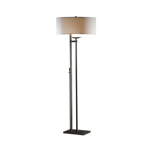 A modern Hubbardton Forge Rook Floor Lamp features a sleek black base and a tall, slender pole, topped with a large, round, cream-colored lampshade. The design is simple and elegant.