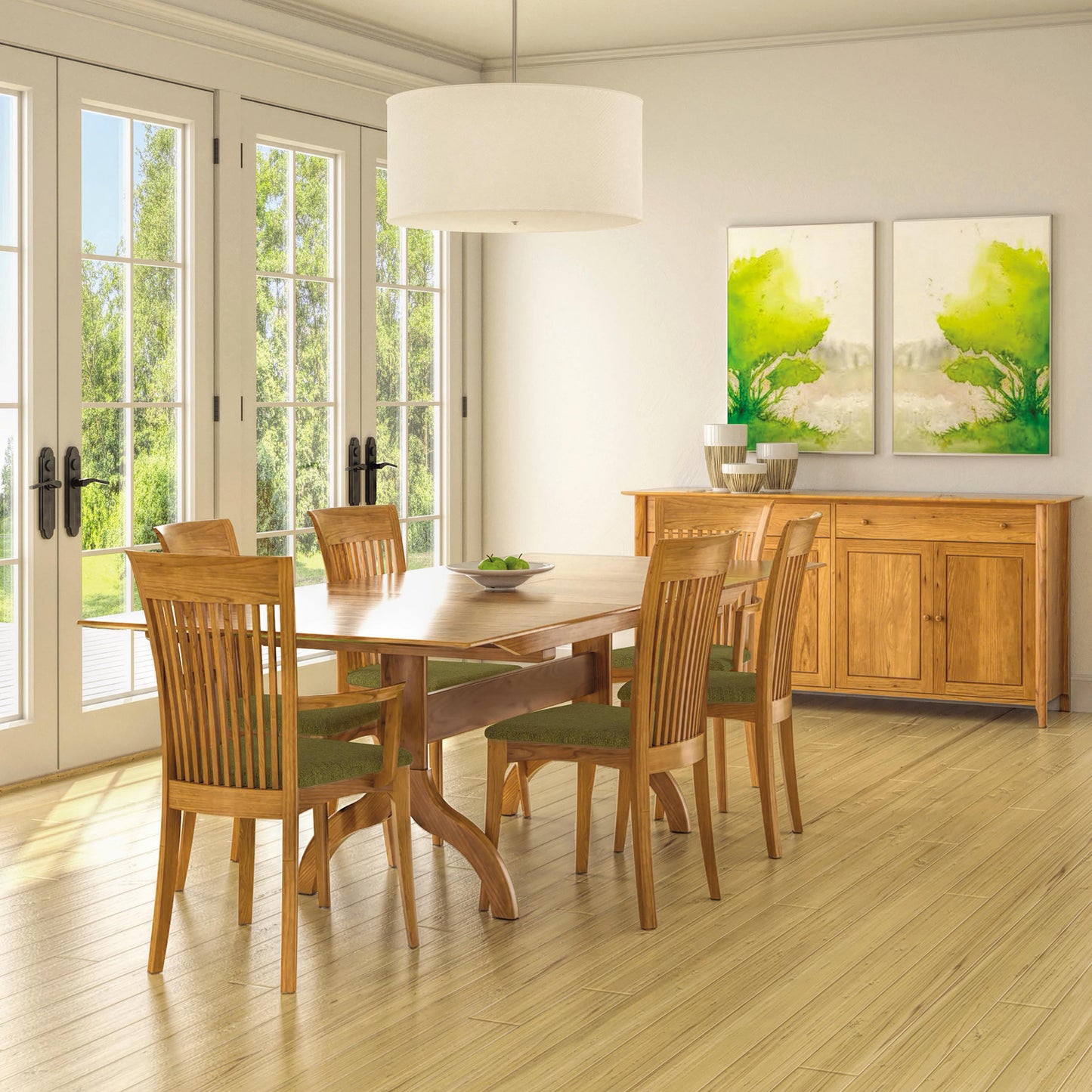 A dining room with a Sarah Shaker Trestle Extension Table by Copeland Furniture and six matching wooden chairs with green cushions. The room features large glass doors, two green and white abstract paintings on the walls, and a wooden sideboard adorned with decorative items. A large pendant light hangs above the Shaker dining room collection’s table.