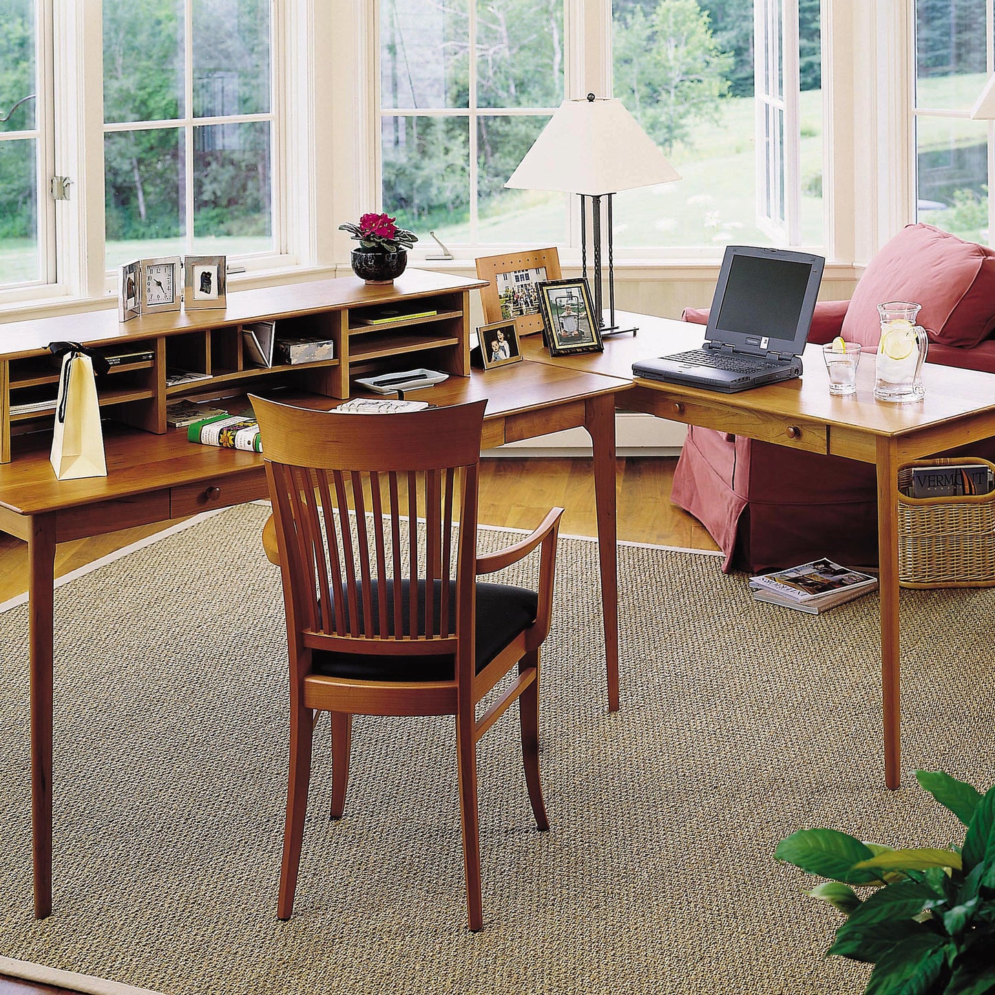 The image shows a home office setup with a Sarah Large Desk by Copeland Furniture, featuring an L-shaped Shaker design. On the desk are a laptop, lamp, framed photos, a potted plant, and office supplies. There is a wooden chair with a black cushion, and the room has large windows letting in natural light.