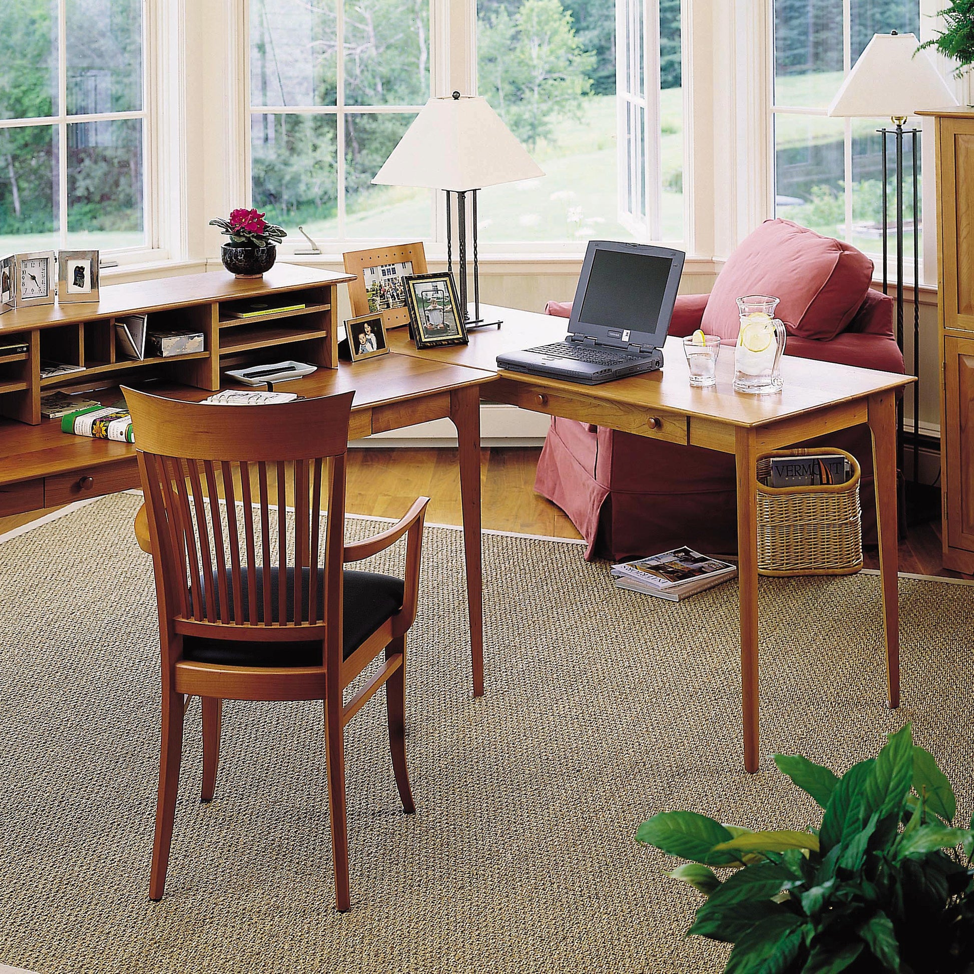 A home office with a large window and a view of the Vermont greenery outside. The Sarah Return with Keyboard Tray by Copeland Furniture features a laptop, a glass of water, and a jug on its wooden surface. A matching wooden chair is placed at the desk, while a pink armchair sits nearby. Shelving units hold books, framed photos, and decorative items curated by Sarah Shaker.