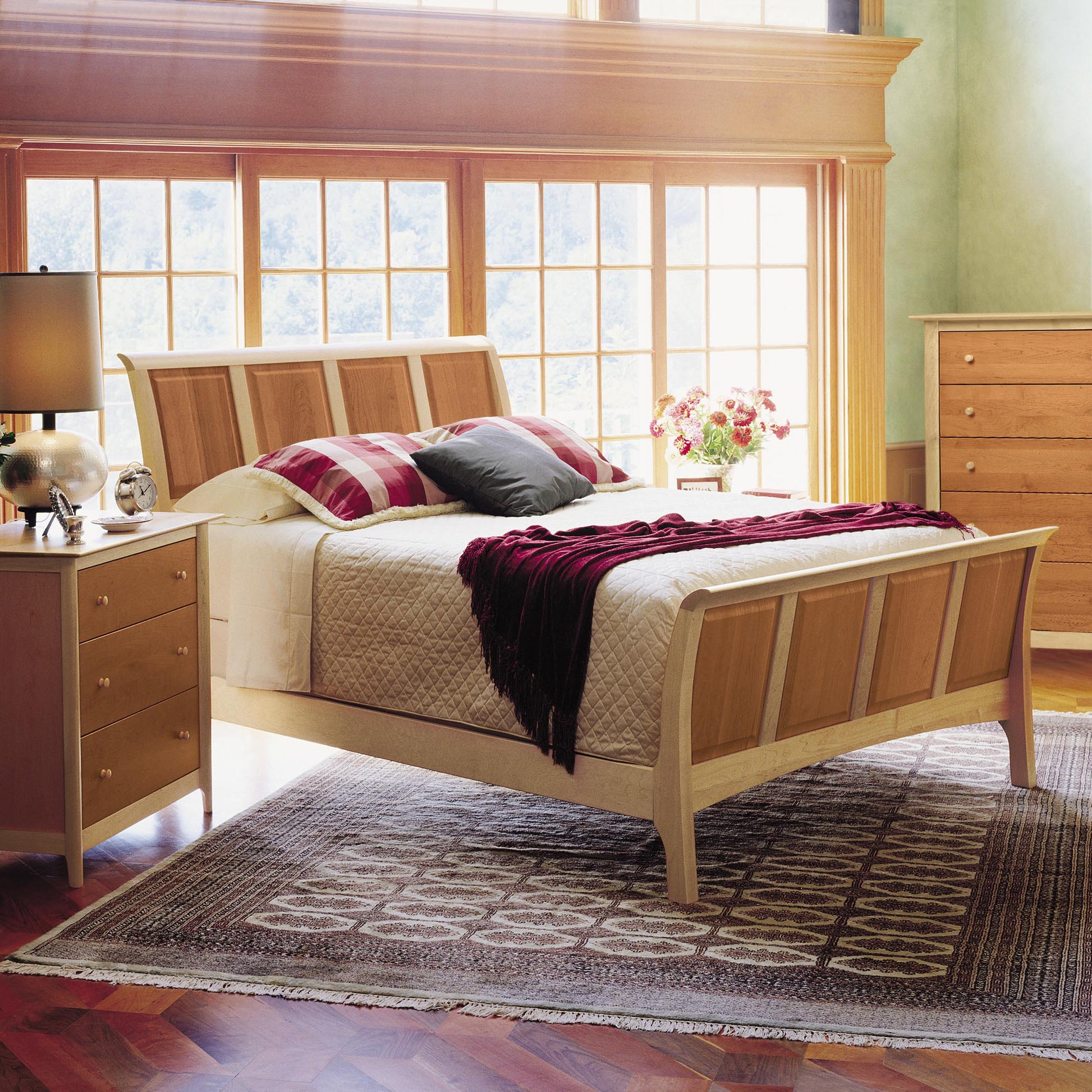 A bedroom with a luxury Sarah High Footboard Sleigh Bed from Copeland Furniture's Shaker-style Sarah Bedroom Furniture Collection, featuring a quilted white bedspread. Pillows in red and gray are on the bed along with a maroon throw. The room has a bedside table with a lamp, an eco-friendly cherry wood dresser, and large windows letting in natural light.