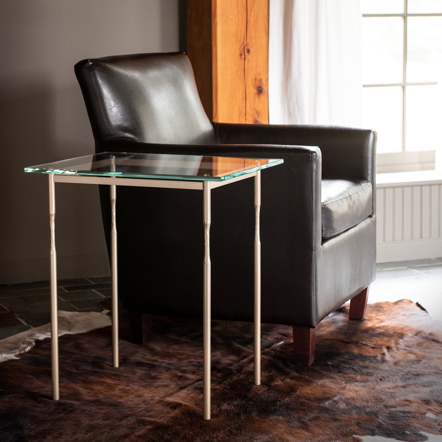 The modern home is beautifully enhanced with the elegant addition of the Hubbardton Forge Senza Side Table, featuring a sleek glass top and complemented by a leather chair of fine tailoring.
