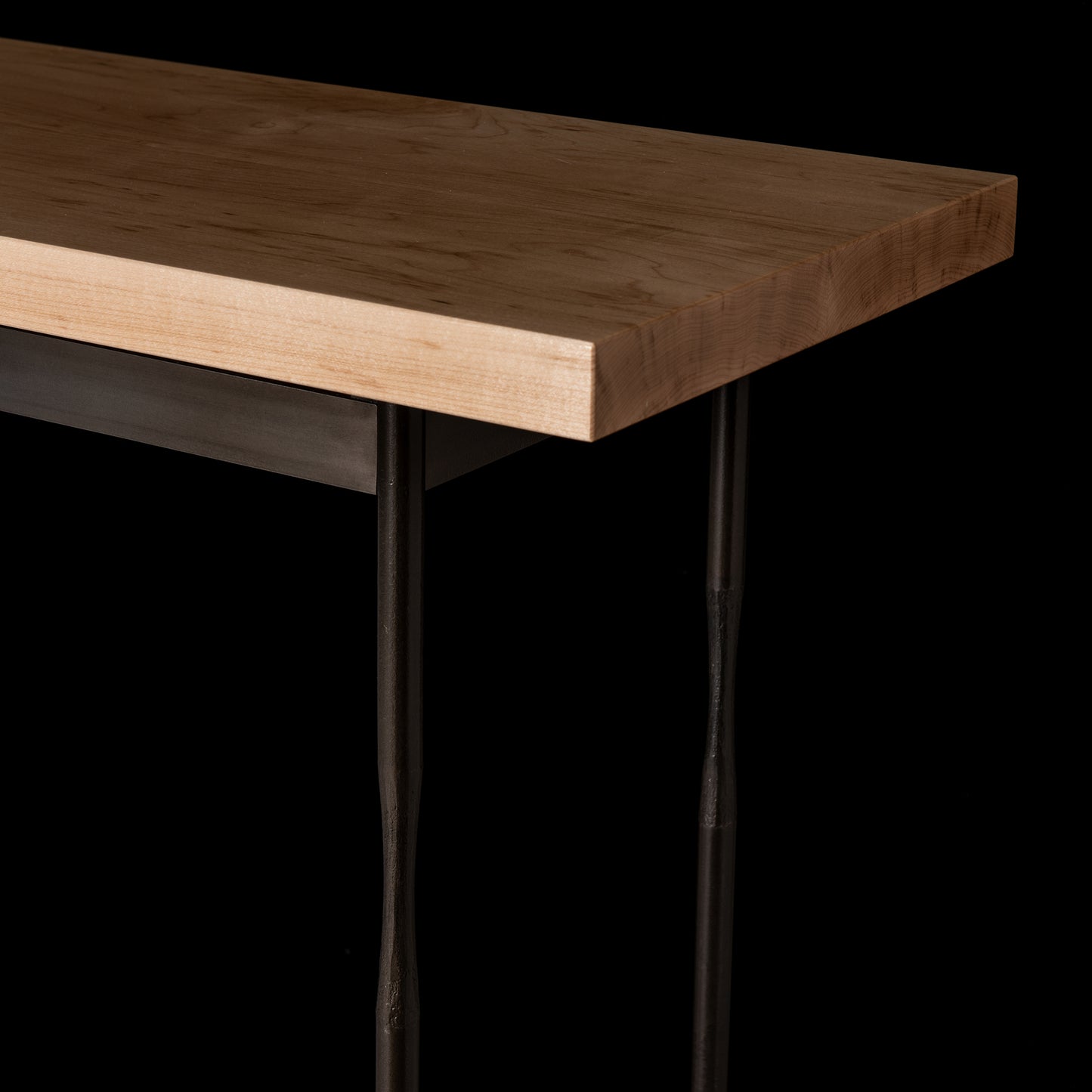 In the image, a Hubbardton Forge Senza Wood Top Console Table is depicted with a handcrafted steel leg supporting its Vermont maple wood top. The table features a light brown, rectangular tabletop with slim black metal legs against a black background, beautifully highlighting the exquisite natural grain pattern of the wood surface.