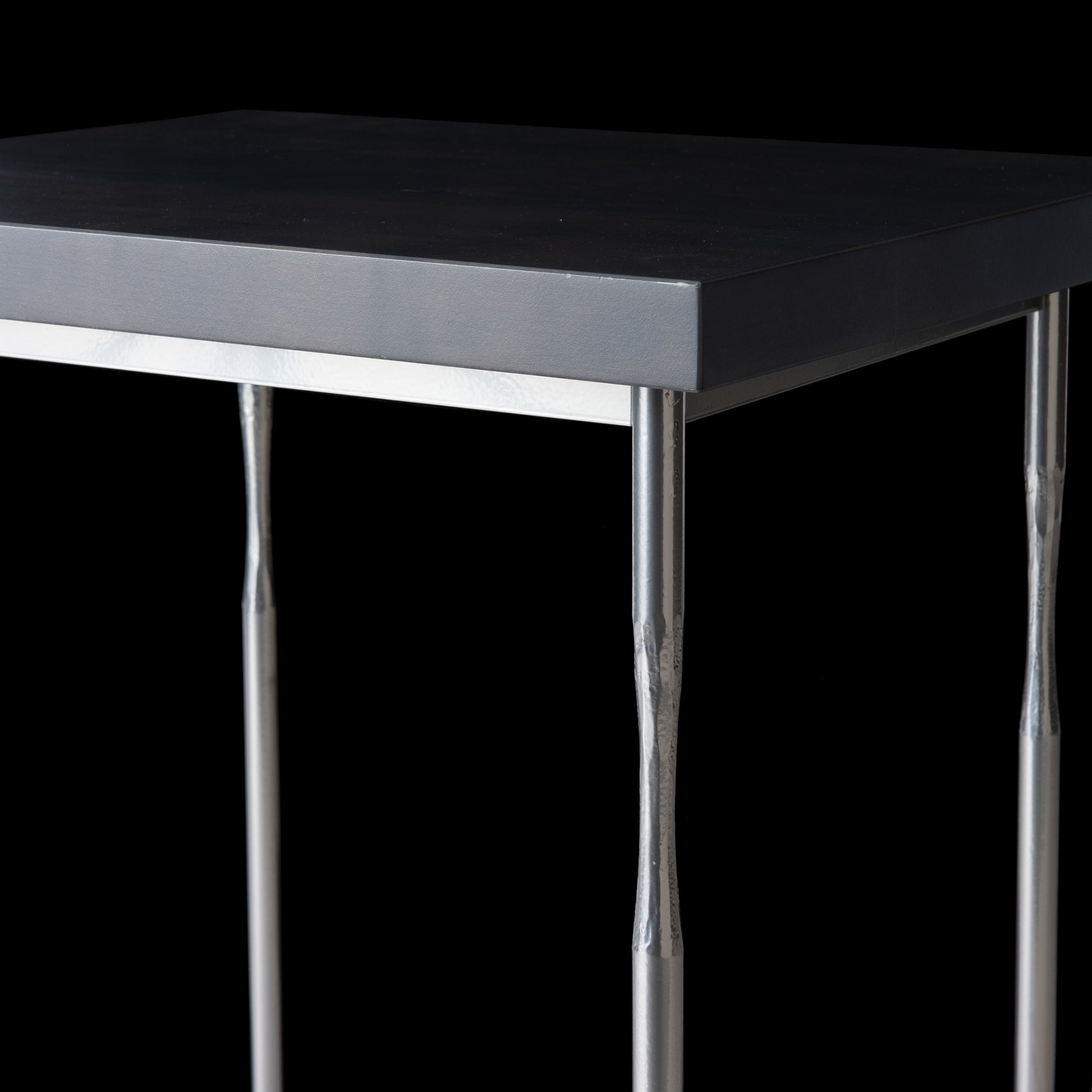 A close-up view of the elegant Senza Wood Top Side Table by Hubbardton Forge showcases a black rectangular top and four slim metal legs, set against a black background.