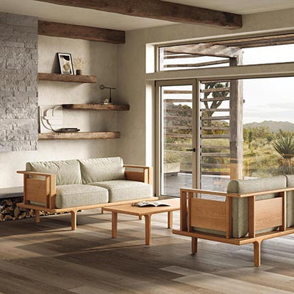 Modern living room with wooden furniture including a couch, armchair, and coffee table. A stone accent wall features wooden shelves with decor items. Large windows offer a view of a scenic landscape with mountains and greenery.