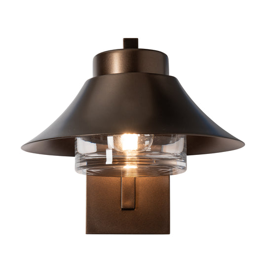 Stowe Outdoor Sconce in dark bronze, featuring a hand-forged steel frame, wide metal shade, and a clear ribbed glass cylinder with a lit bulb.