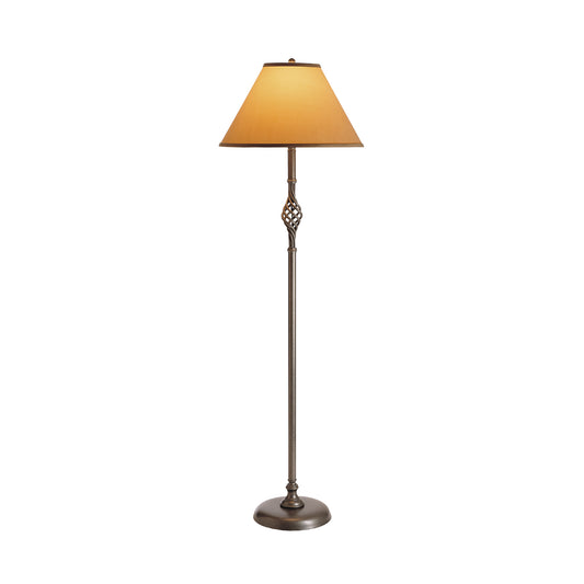 A tall Twist Basket Floor Lamp by Hubbardton Forge with an ornate metal base and a slender pole, supporting a large, bell-shaped lampshade in a soft amber color. The hand-crafted lamp is isolated on a white background.