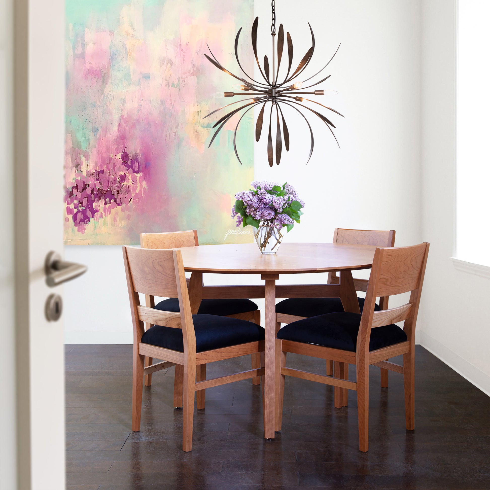 A small dining area showcases a round Vermont Modern Round Solid Top Pedestal Table by Lyndon Furniture, crafted from solid organic wood. The table is surrounded by four matching chairs with dark cushions. Above, a modern pendant light illuminates the space. A colorful abstract painting adorns the white wall, and a vase of flowers sits elegantly on the table.