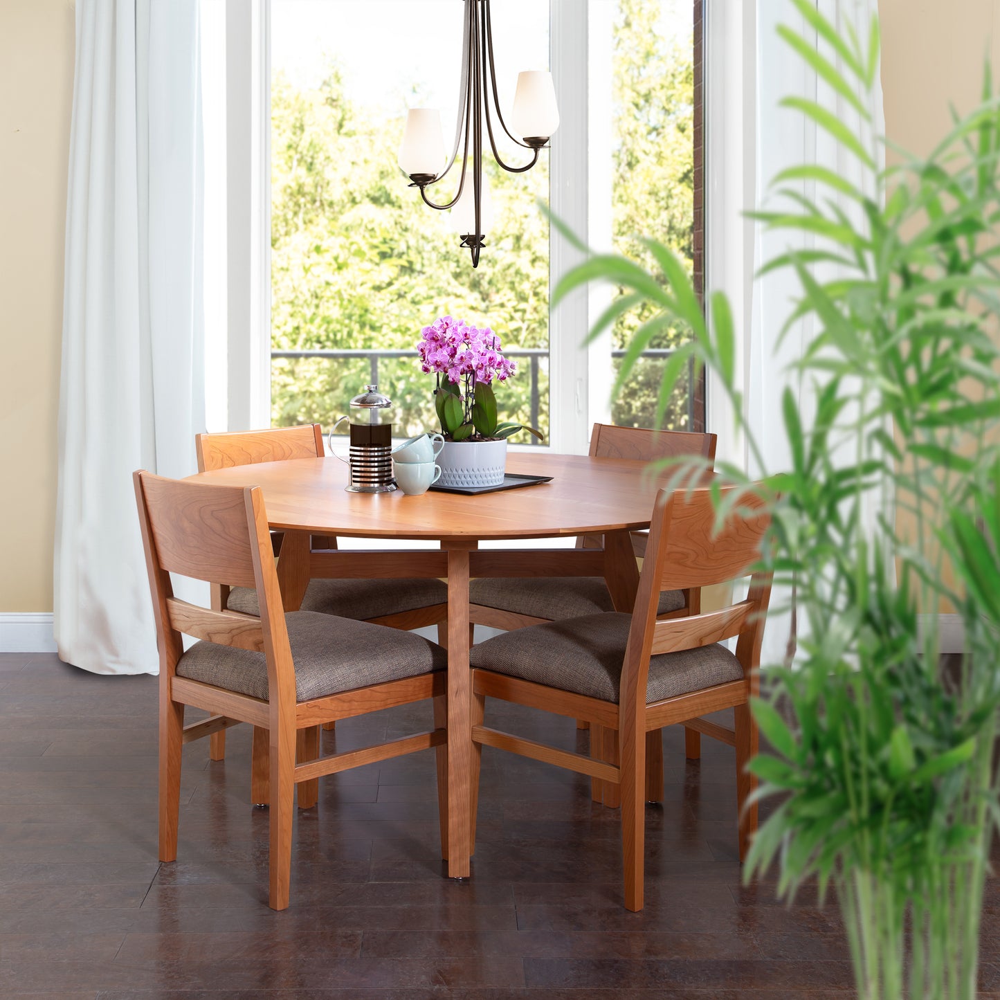 A round Vermont Modern Round Solid Top Pedestal Table by Lyndon Furniture, paired with four matching chairs, graces a bright room with large windows. A potted purple orchid and various items adorn the table. A hanging chandelier provides lighting, and a green potted plant is partially visible in the foreground, highlighting the handmade furniture from Vermont.