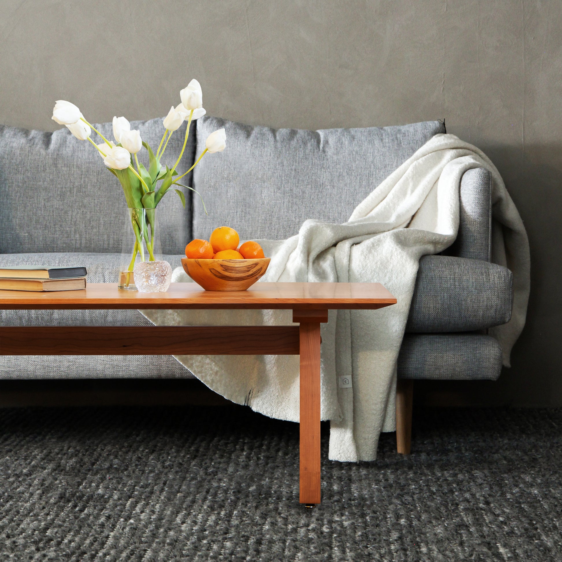 Gray sofa adorned with white blanket; Lyndon Furniture Vermont Modern Trestle Coffee Table featuring vase of tulips and fresh oranges.