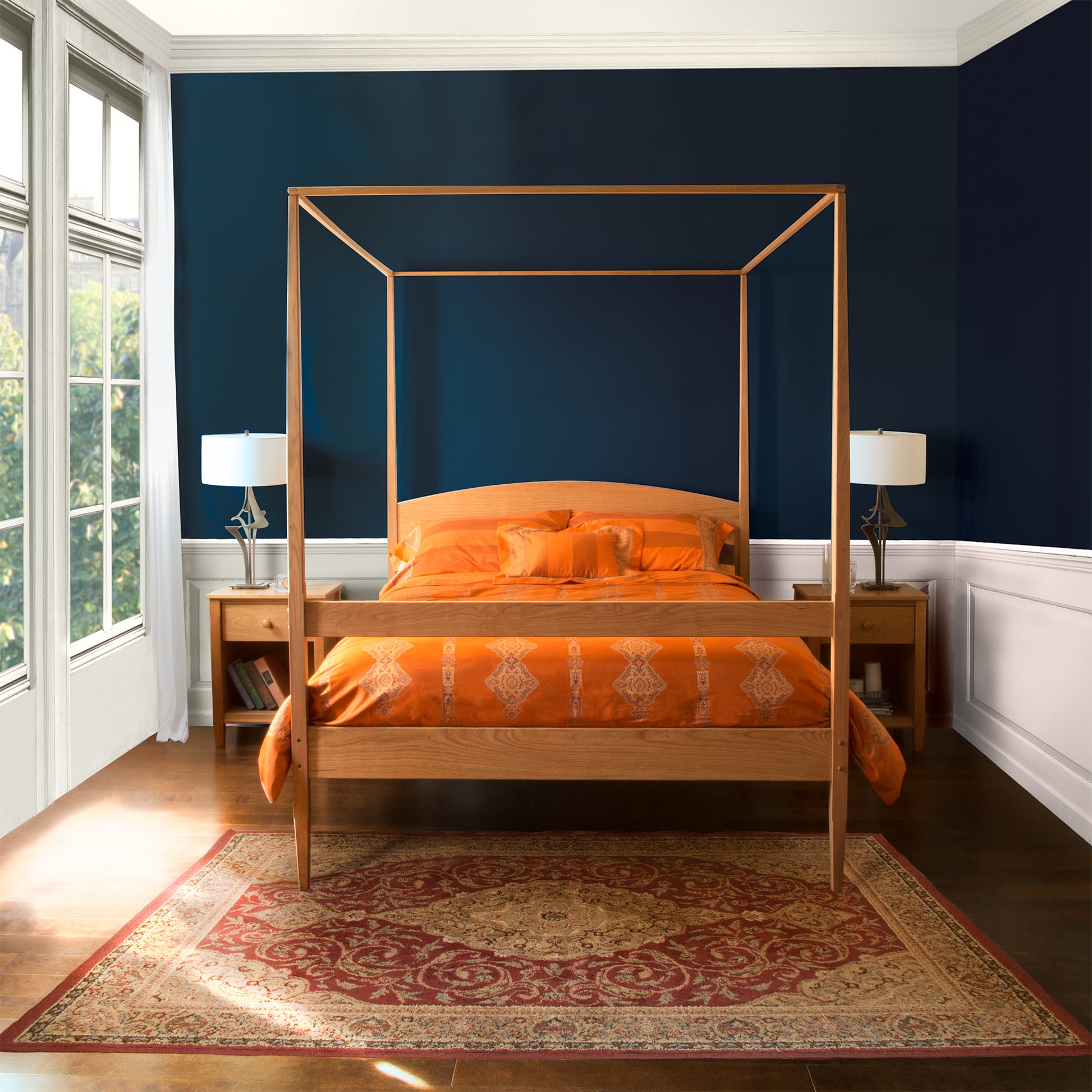 A bedroom showcasing a Vermont Shaker Four Poster Bed with Canopy from Maple Corner Woodworks, adorned with orange bedding and pillows. On either side of the bed stand two matching wooden nightstands topped with lamps. The floor is enhanced by a patterned red area rug, while large windows to the left illuminate the room with natural light.