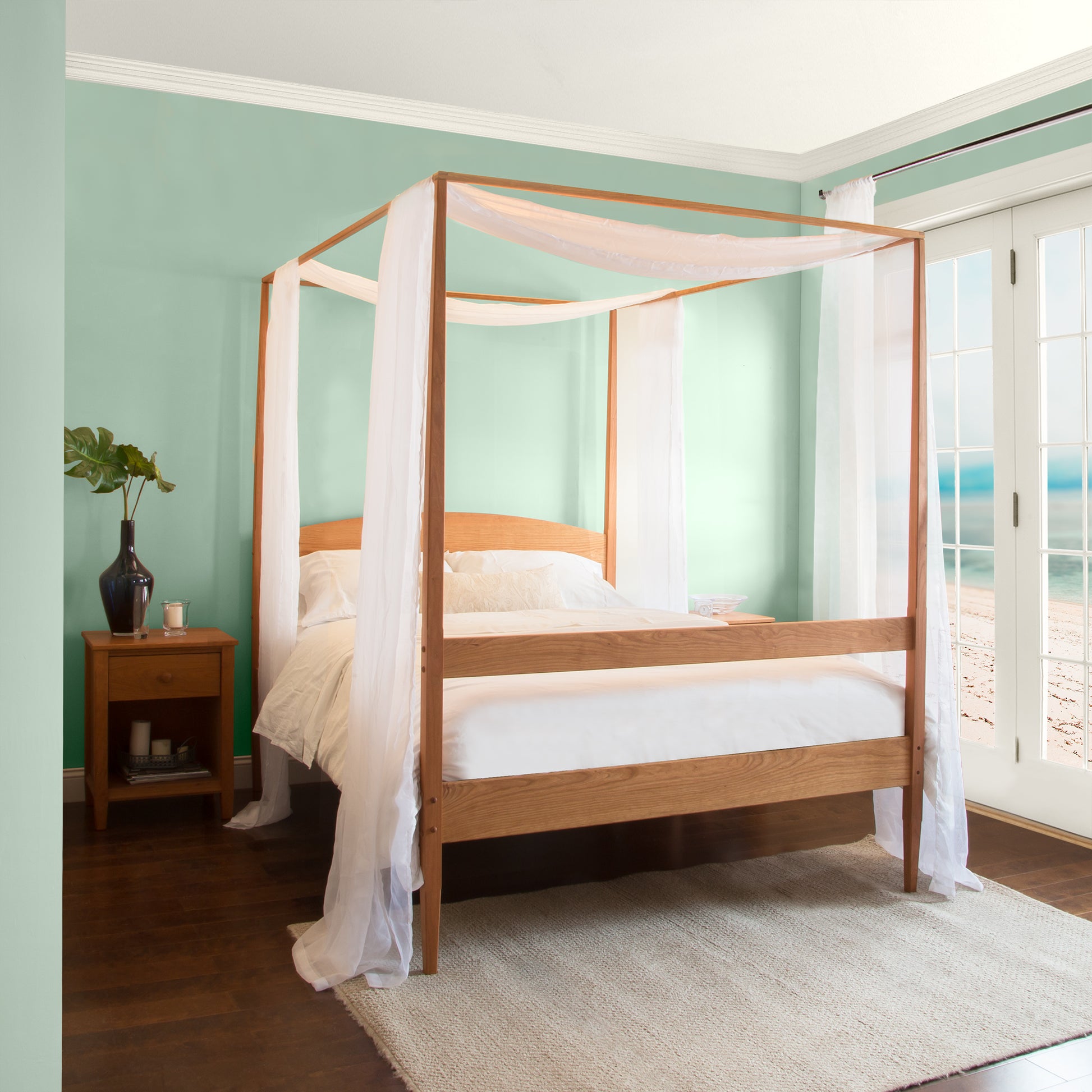 A serene bedroom featuring a Maple Corner Woodworks Vermont Shaker Four Poster Bed with Canopy draped with sheer white curtains, light teal walls, dark wooden flooring, and a small wooden side table with a plant. Large windows let