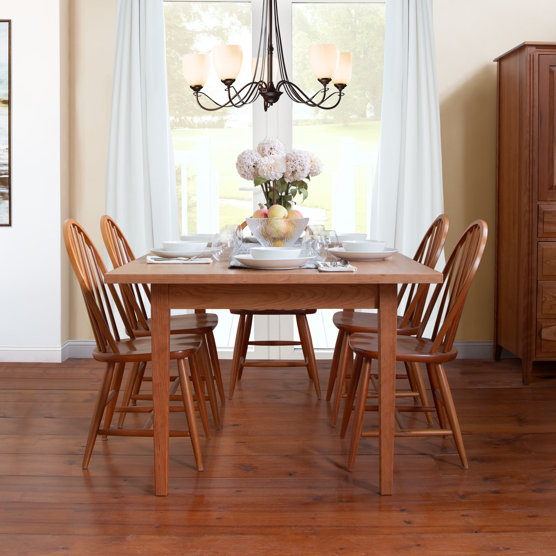 A Vermont Shaker Solid Top Harvest Table from Maple Corner Woodworks in natural cherry wood is teamed with six chairs, situated in a well-lit room with expansive windows. The table is elegantly set with white tableware and a centerpiece displaying white flowers and fruit, beneath a black chandelier on hardwood floors.