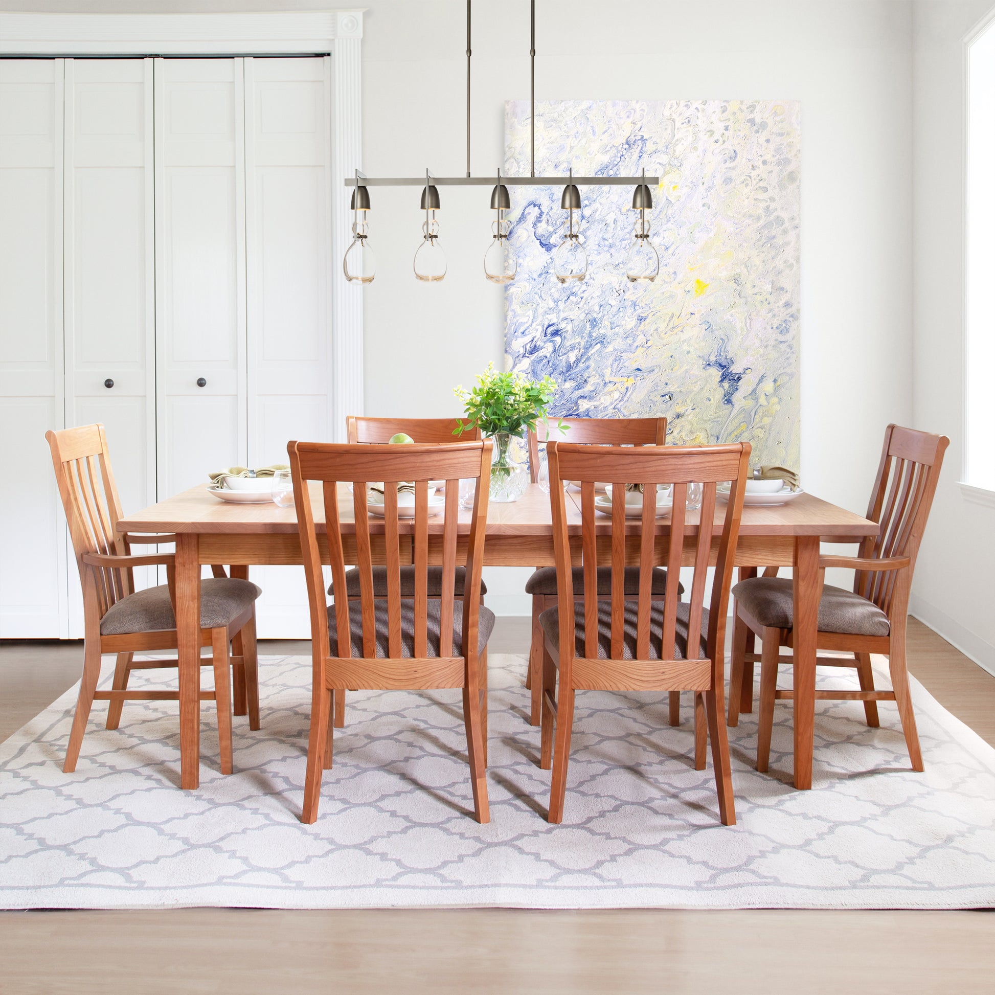 A dining room showcases the Vermont Shaker Rectangular Extension Dining Table by Maple Corner Woodworks, accompanied by six matching chairs on a patterned rug. A modern chandelier hangs above, casting light over the elegantly set table adorned with plates and a vase of flowers. Light-colored walls feature an abstract painting.