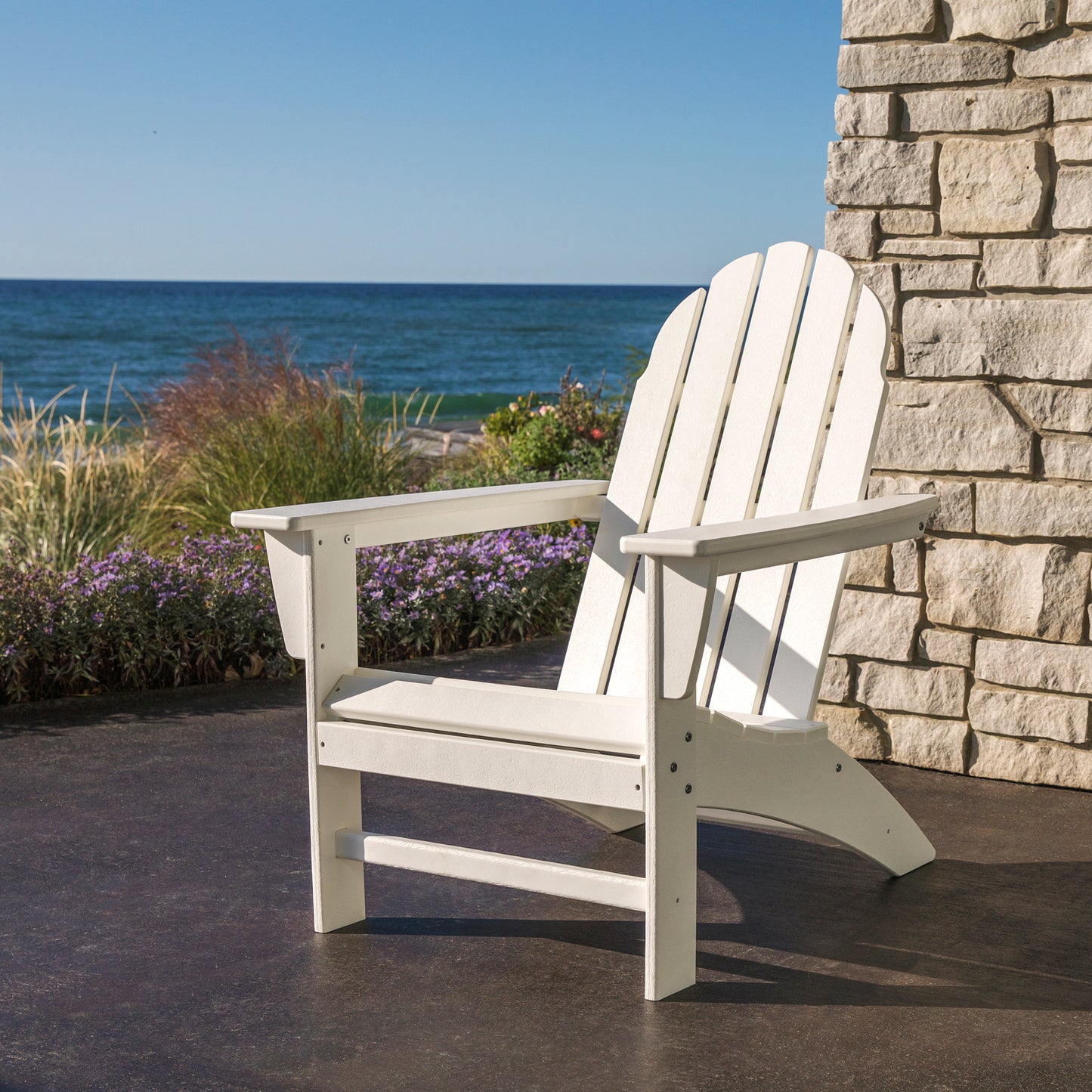 A POLYWOOD Vineyard Adirondack Chair, made from sturdy POLYWOOD® lumber, sits elegantly on a stone patio with an ocean view. A stone wall adds to the scene's charm, while grasses and flowers decorate the patio's edge beneath a clear blue sky.