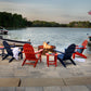 A serene patio scene features four POLYWOOD Vineyard Adirondack Chairs, crafted from durable POLYWOOD® lumber, arranged around a round fire pit by the lakeside. Two of the chairs are red and two are blue. A small table nearby holds snacks and drinks. A boat is docked close to shore, with trees encircling the lake under a cloudy sky.