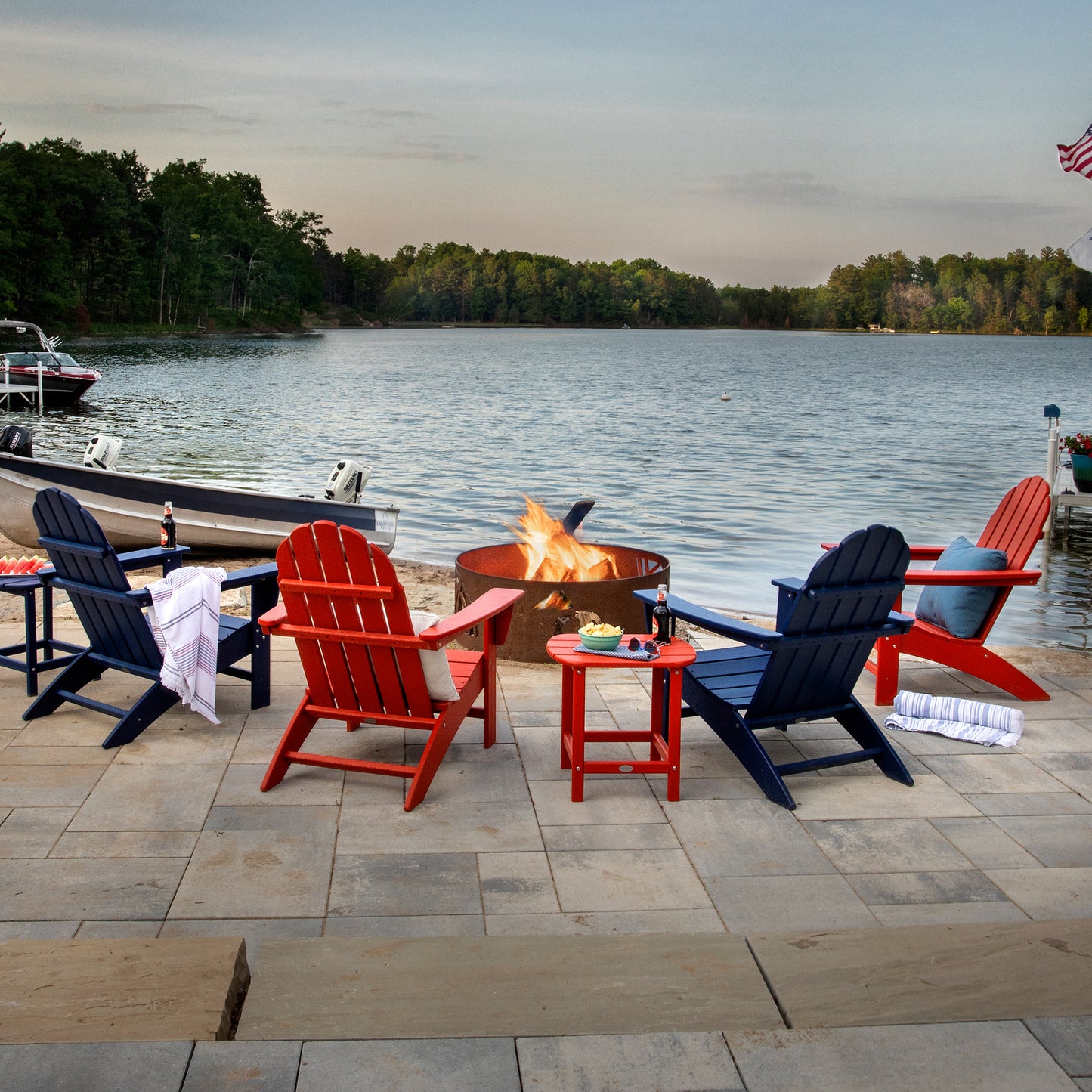 A serene patio scene features four POLYWOOD Vineyard Adirondack Chairs, crafted from durable POLYWOOD® lumber, arranged around a round fire pit by the lakeside. Two of the chairs are red and two are blue. A small table nearby holds snacks and drinks. A boat is docked close to shore, with trees encircling the lake under a cloudy sky.