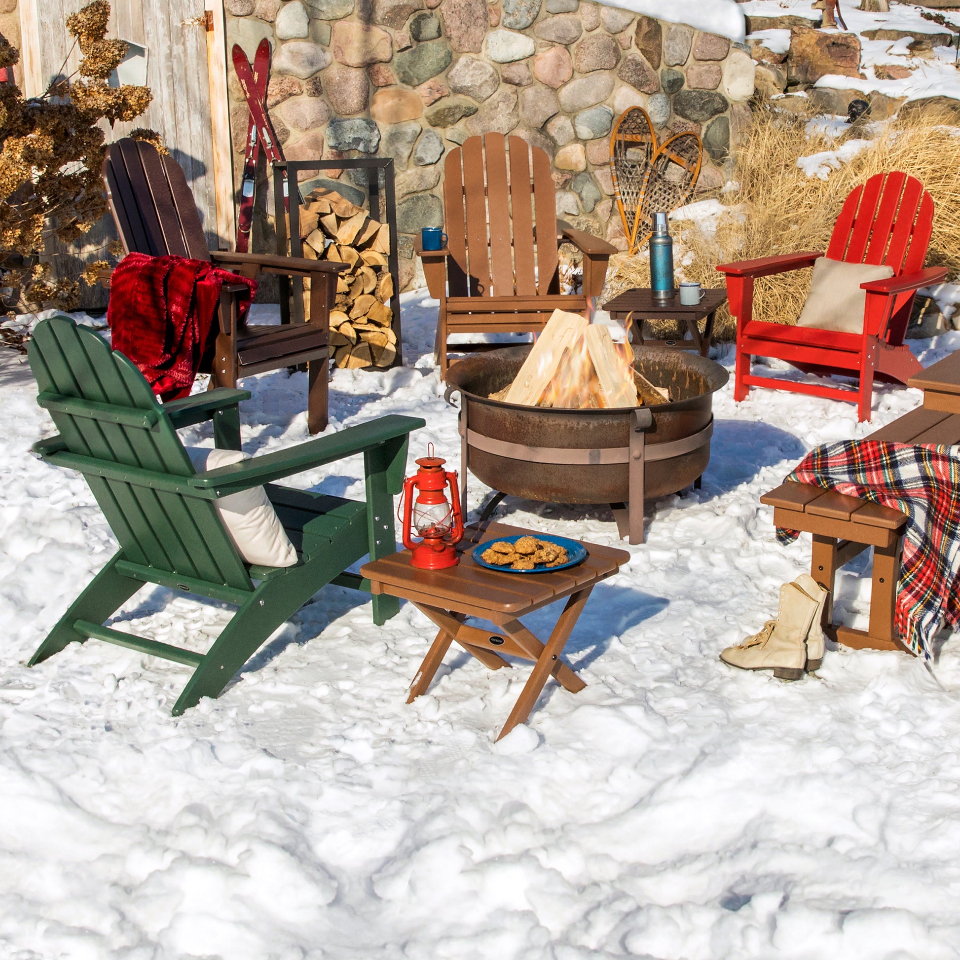 In a snowy outdoor scene, a round fire pit is encircled by POLYWOOD Vineyard Adirondack Chairs, crafted from durable POLYWOOD lumber. A nearby small table showcases a red lantern and a plate of cookies. In the background, there's a stack of firewood and snowshoes, while blankets and pillows enhance the coziness of this delightful outdoor furniture arrangement.