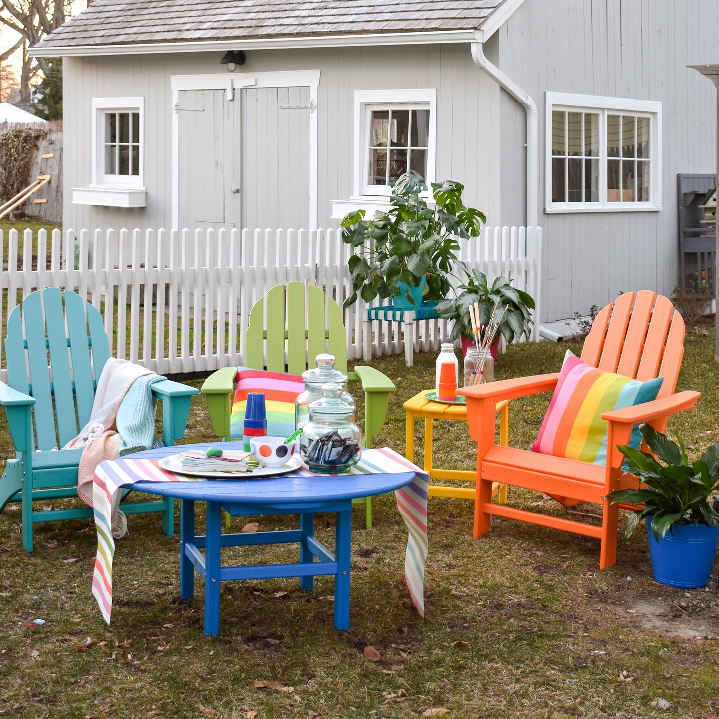 In a backyard setting, vibrant POLYWOOD Vineyard Adirondack Chairs in shades of blue, green, yellow, and orange are arranged around a blue table covered with a striped cloth. The chairs are made from durable POLYWOOD lumber and beautifully offset the gray shed and white picket fence. A plant and jar adorn the tabletop.