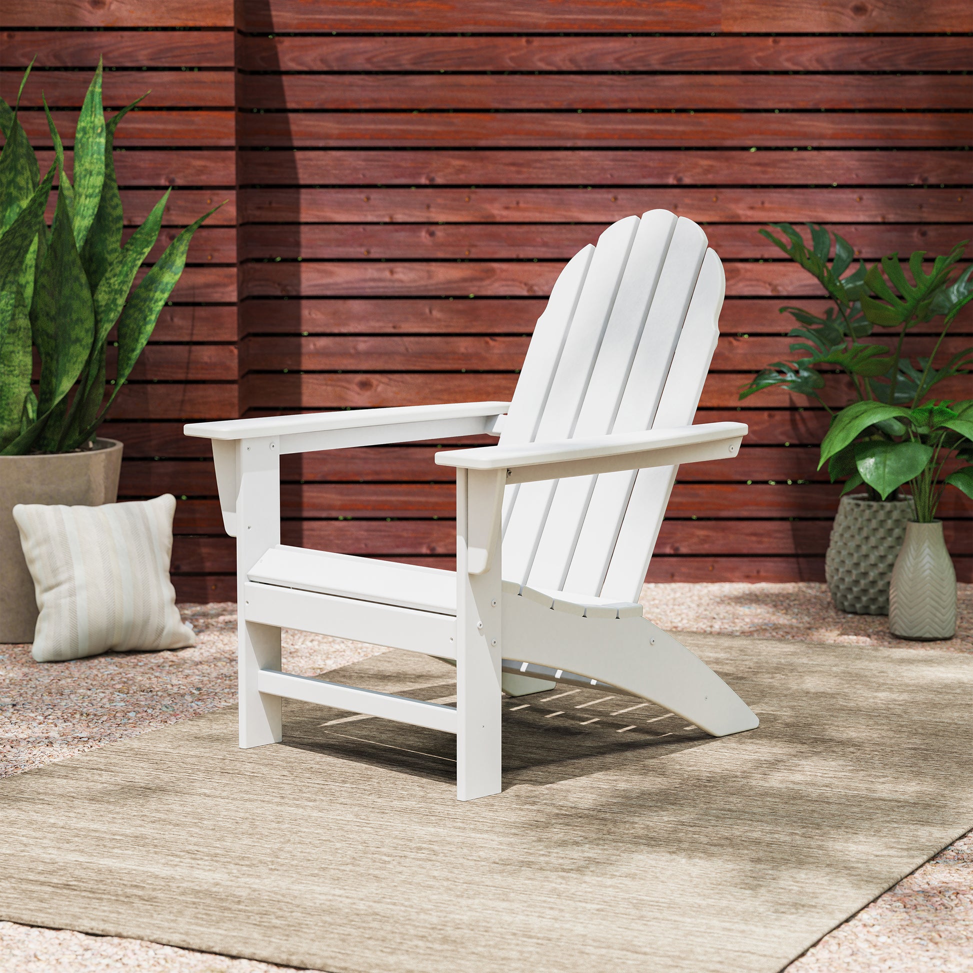 A white POLYWOOD Vineyard Adirondack Chair, meticulously crafted from sturdy POLYWOOD® lumber, is elegantly placed on a beige rug surrounded by vibrant potted plants and complemented by a striped cushion. The outdoor furniture piece is bordered by a wooden fence with horizontal slats, creating a tranquil atmosphere in the patio setting.