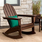 A POLYWOOD Vineyard Adirondack Rocking Chair from the Outdoor Furniture Collection, equipped with a green cushion, rests on a woven rug. It's ideally placed on a porch against a stone wall backdrop, alongside a small side table and decorative grass.