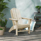 A POLYWOOD Vineyard Curveback Adirondack Chair in weather-resistant beige is positioned on a tiled patio surrounded by potted plants, such as a cactus and a leafy plant. A striped cushion rests on the ground nearby, enhancing the cozy outdoor setting against the light green stucco wall.