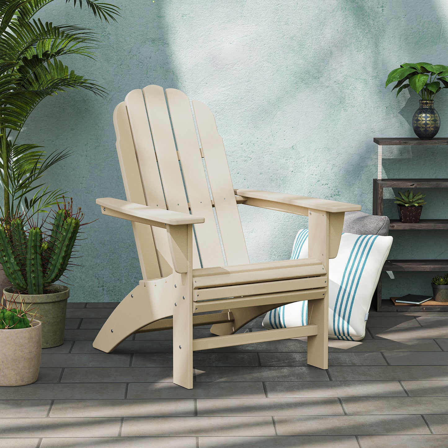 A POLYWOOD Vineyard Curveback Adirondack Chair in weather-resistant beige is positioned on a tiled patio surrounded by potted plants, such as a cactus and a leafy plant. A striped cushion rests on the ground nearby, enhancing the cozy outdoor setting against the light green stucco wall.