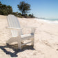 A POLYWOOD Vineyard Curveback Adirondack Chair, known for its weather-resistant outdoor design, sits empty on a sandy beach with the ocean visible to the right. A clear blue sky and a few trees are in the background, enhancing the serene and bright setting.