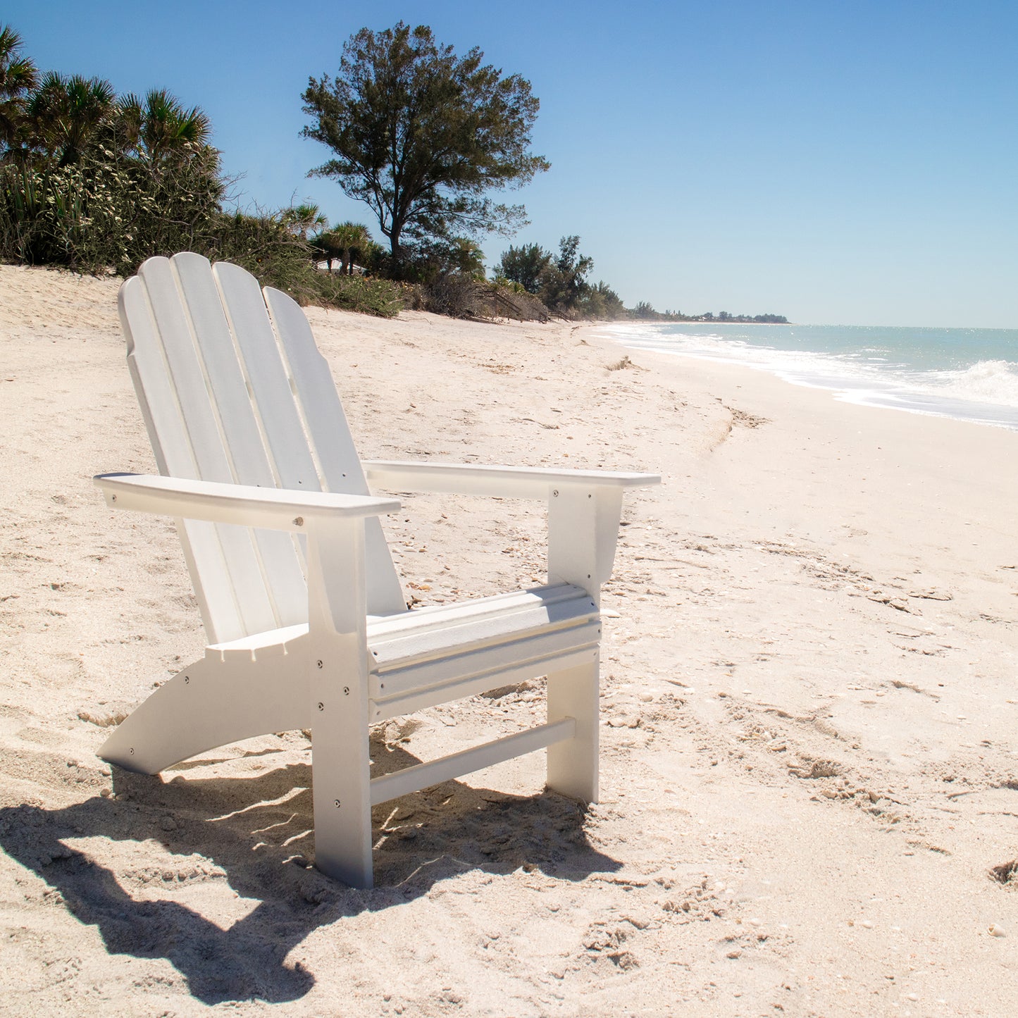 A POLYWOOD Vineyard Curveback Adirondack Chair, known for its weather-resistant outdoor design, sits empty on a sandy beach with the ocean visible to the right. A clear blue sky and a few trees are in the background, enhancing the serene and bright setting.