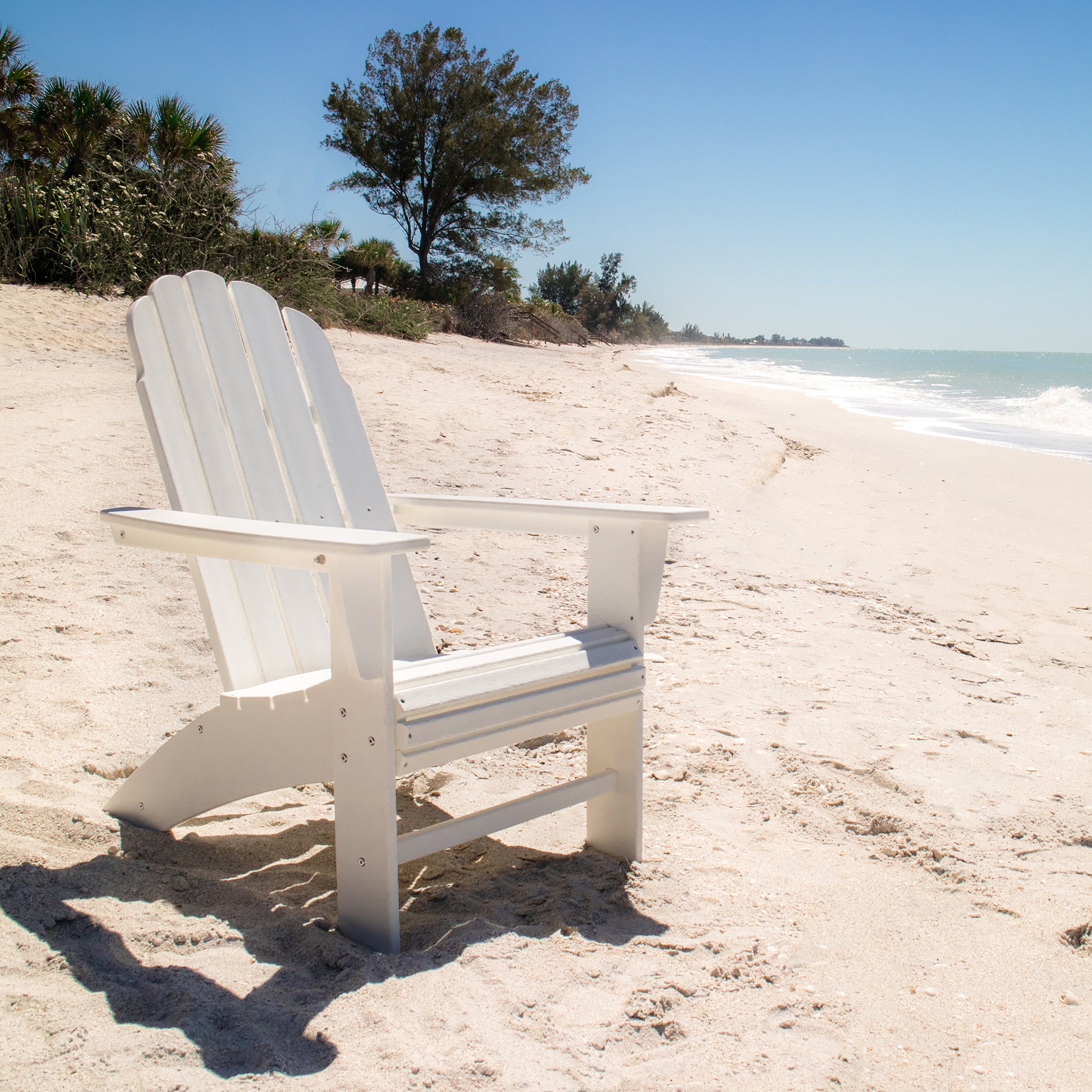 A POLYWOOD Vineyard Curveback Adirondack Chair, known for its weather-resistant outdoor design, sits empty on a sandy beach with the ocean visible to the right. A clear blue sky and a few trees are in the background, enhancing the serene and bright setting.