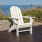 A POLYWOOD Vineyard Curveback Adirondack Chair, made from weather-resistant materials, is placed on a patio overlooking a body of water with a lighthouse in the distance. Tall grasses and large rocks surround the area, and the sky is clear.