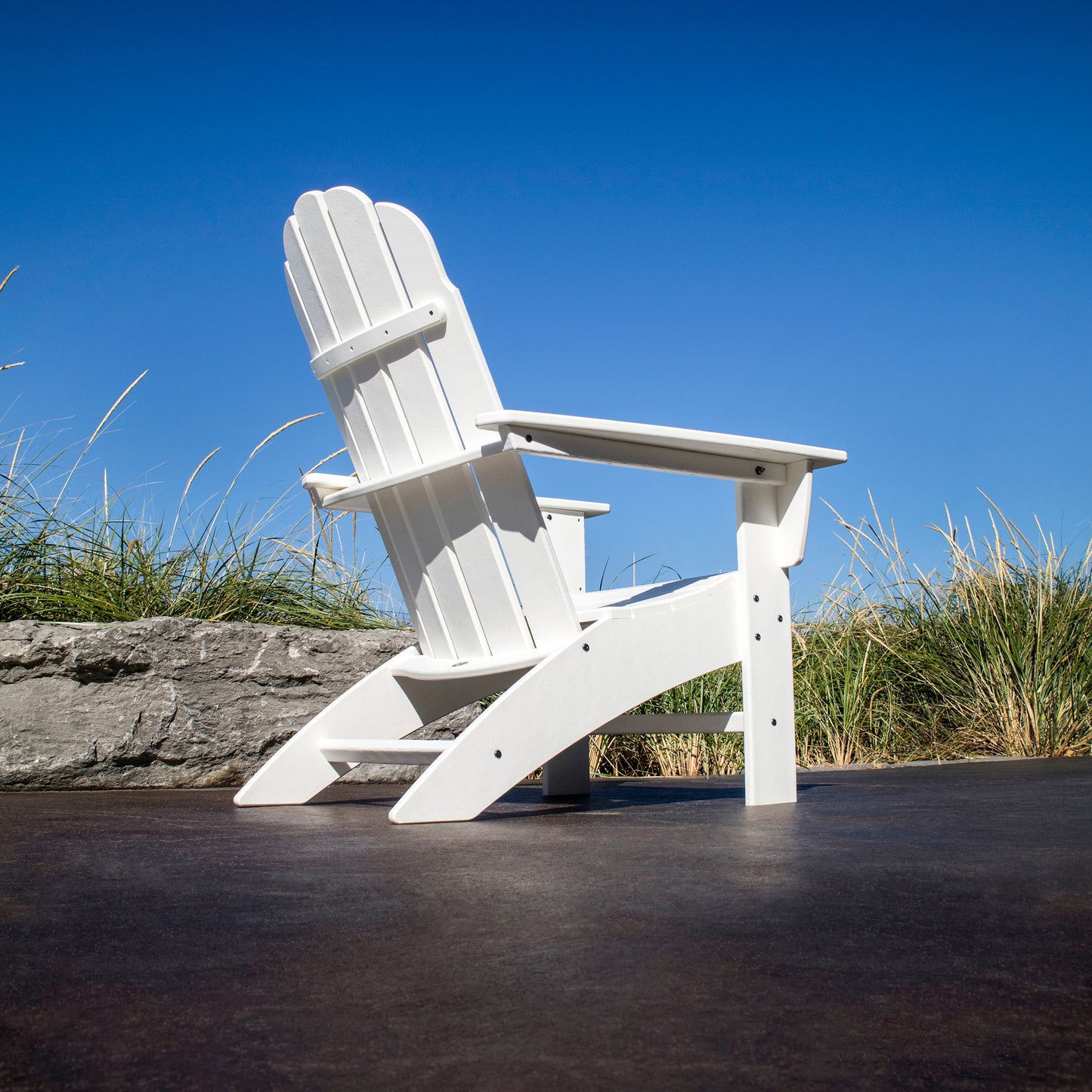 The POLYWOOD Vineyard Curveback Adirondack Chair, designed with weather-resistant materials, sits elegantly on a dark surface outdoors. It is set against a backdrop of clear blue sky, verdant grassy plants, and a rustic stone wall, embodying the allure of outdoor furniture.