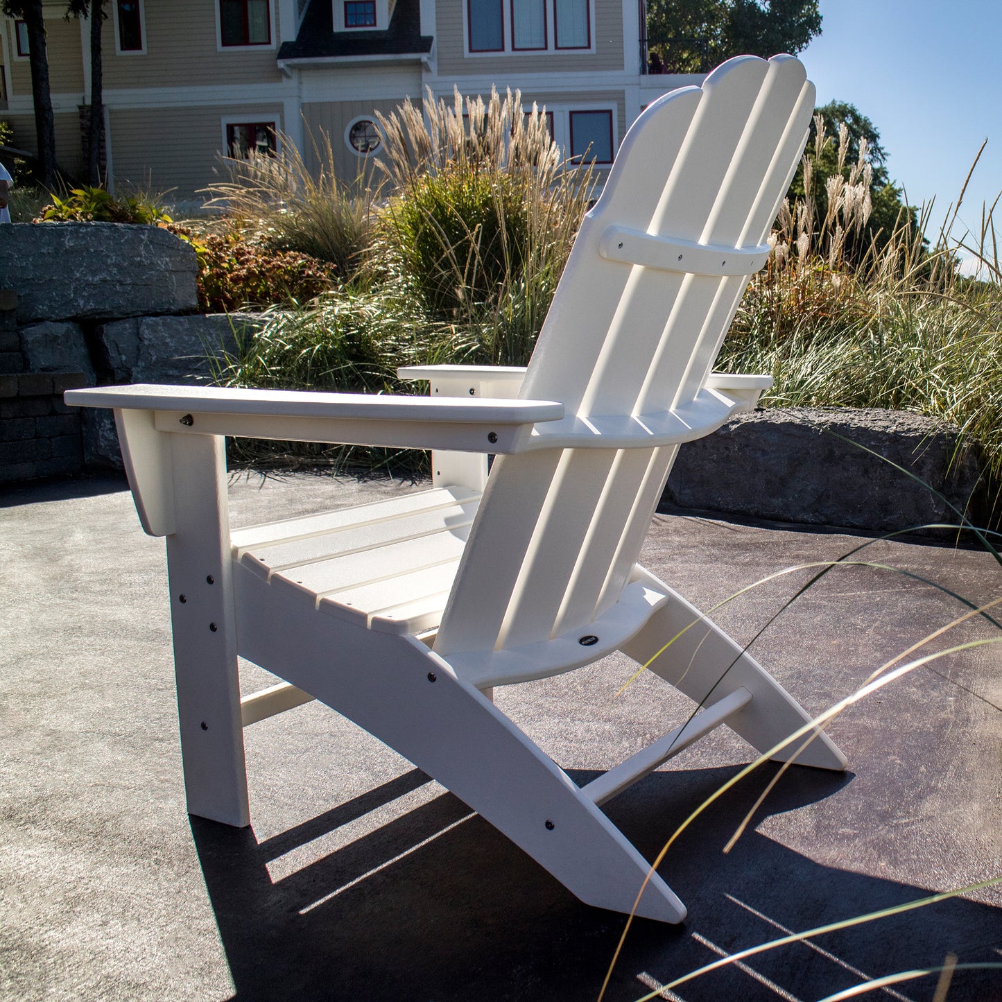 A durable POLYWOOD Vineyard Curveback Adirondack Chair in white rests on a sunlit patio, surrounded by ornamental grasses and stone landscaping. The background displays a partial view of a house with siding and a shingled roof, enhancing the cozy feel of this outdoor furniture setting.