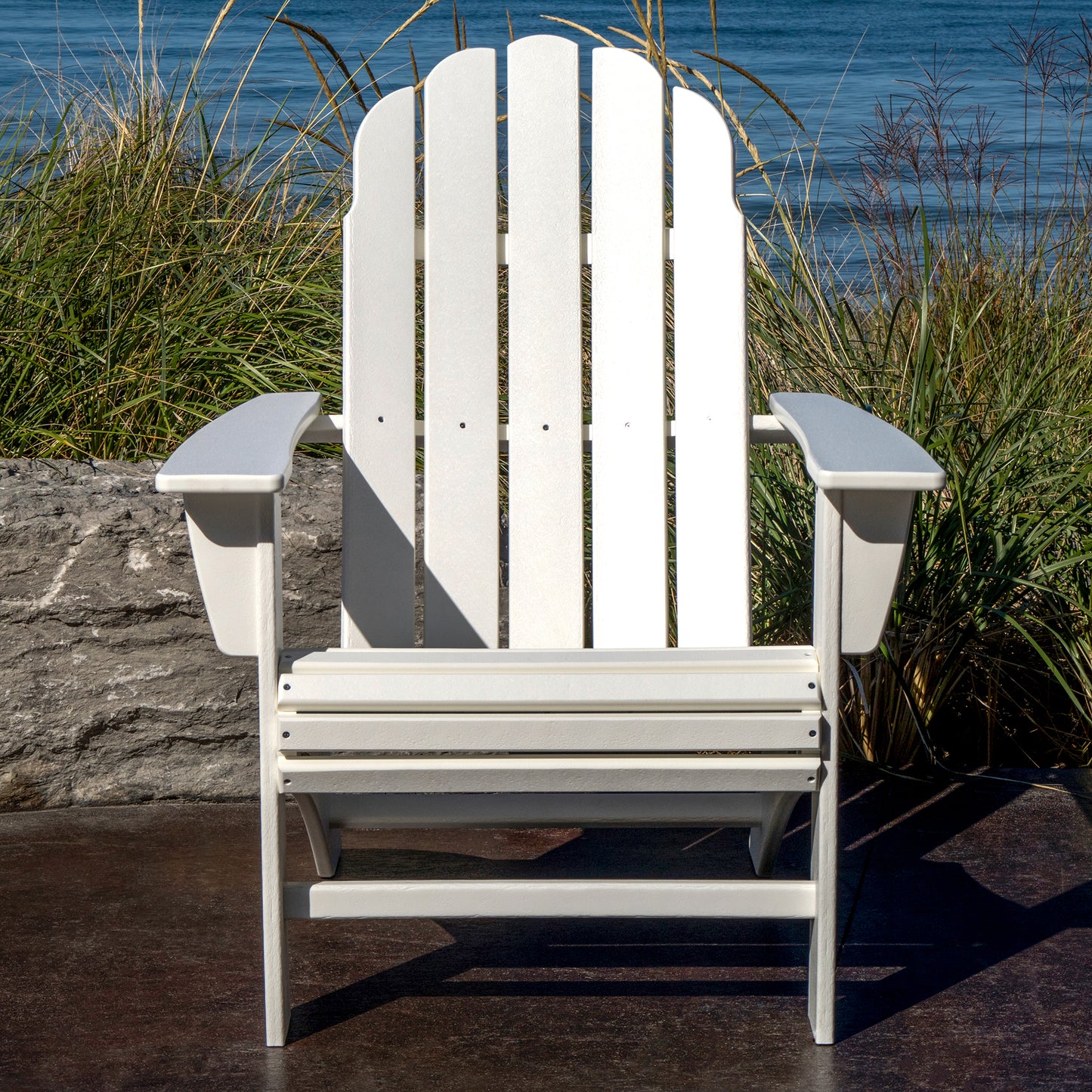 A weather-resistant POLYWOOD Vineyard Curveback Adirondack Chair, in white, sits on a patio with tall grass and a stone wall behind it. Beyond the wall, there is a view of the ocean under a clear blue sky—a perfect piece of outdoor furniture from POLYWOOD for any serene setting.