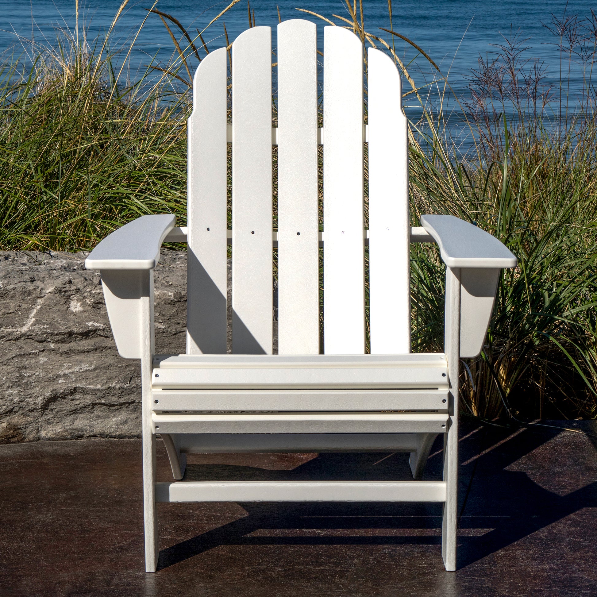 A weather-resistant POLYWOOD Vineyard Curveback Adirondack Chair, in white, sits on a patio with tall grass and a stone wall behind it. Beyond the wall, there is a view of the ocean under a clear blue sky—a perfect piece of outdoor furniture from POLYWOOD for any serene setting.